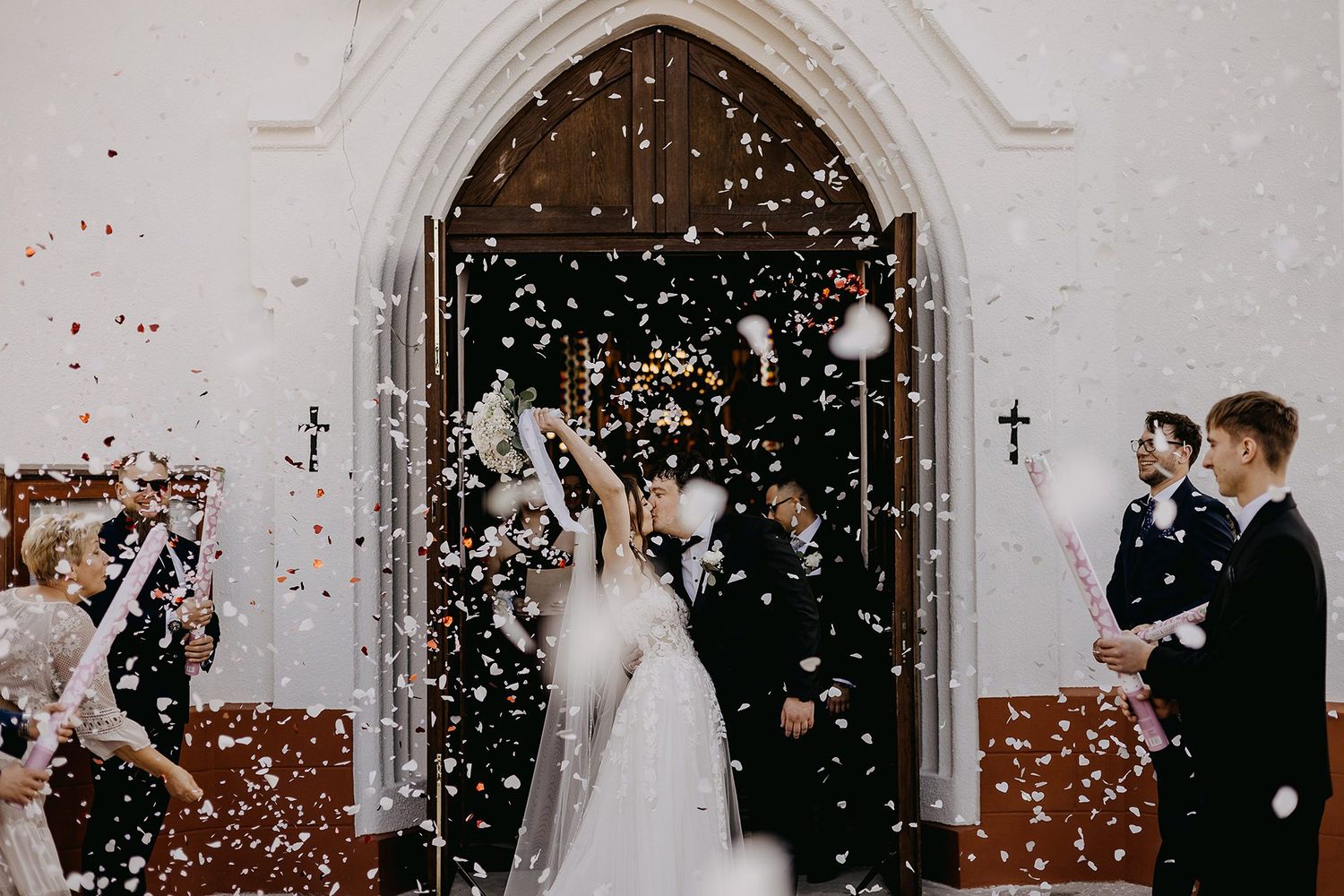 Newlyweds exit a white church amid a shower of colorful confetti, surrounded by joyful wedding guests.
