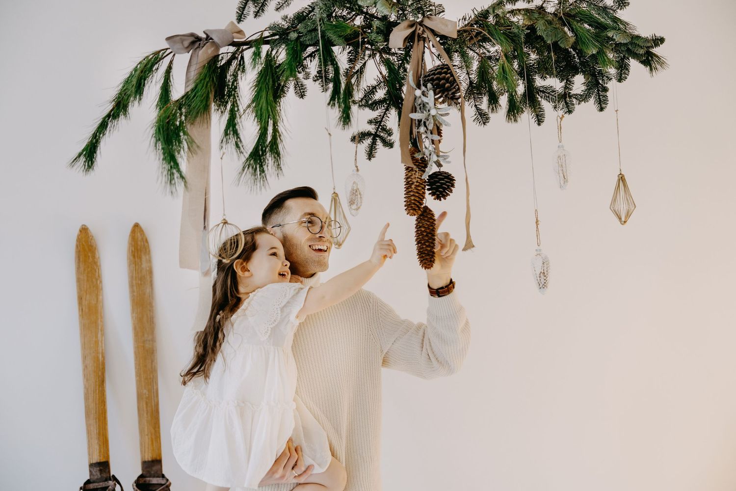 Smiling father holds his young daughter in white dress under a hanging evergreen branch with pinecones, ornaments and vintage wooden skis.