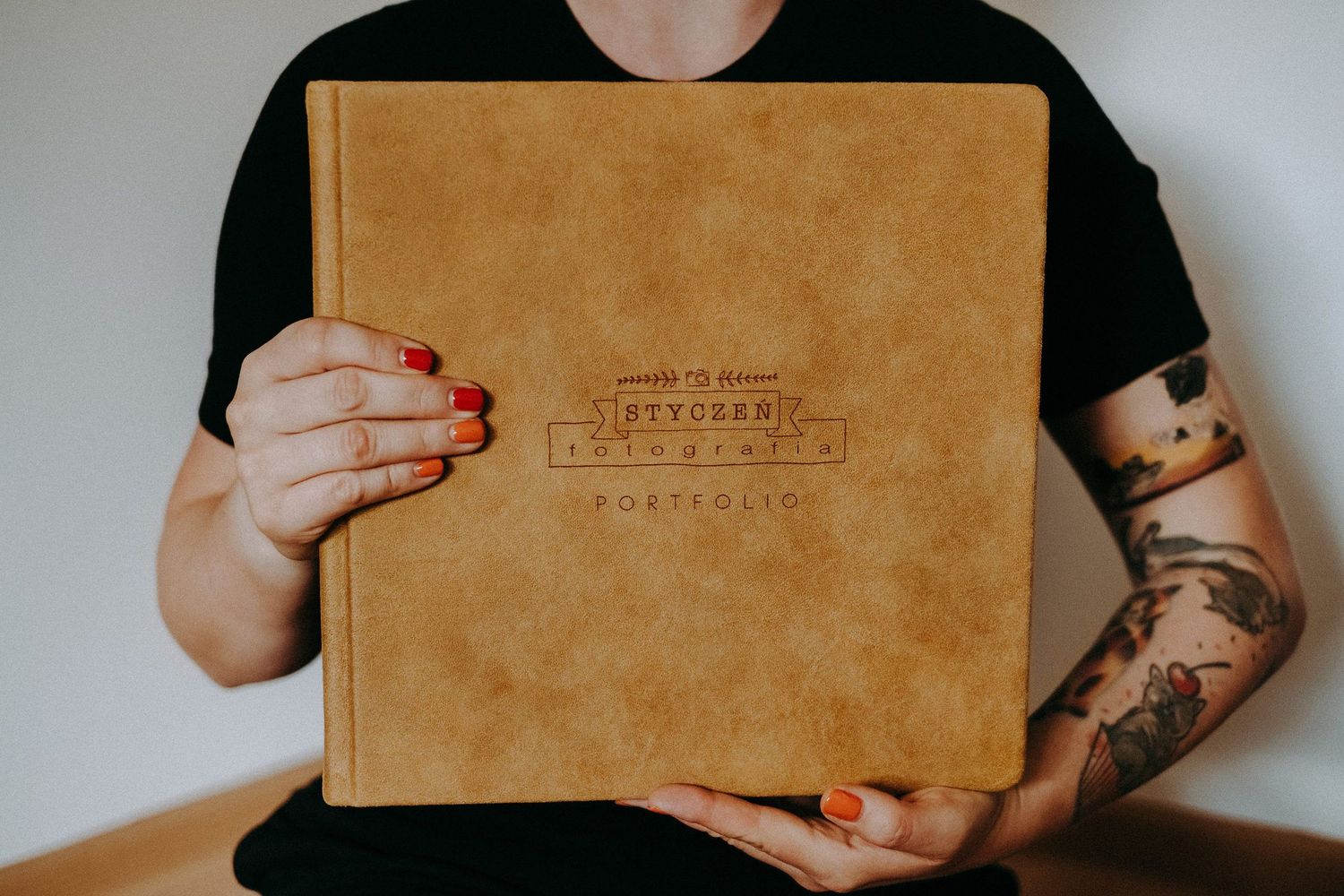 Woman with tattoos and red nails holding a tan leather photography portfolio book.