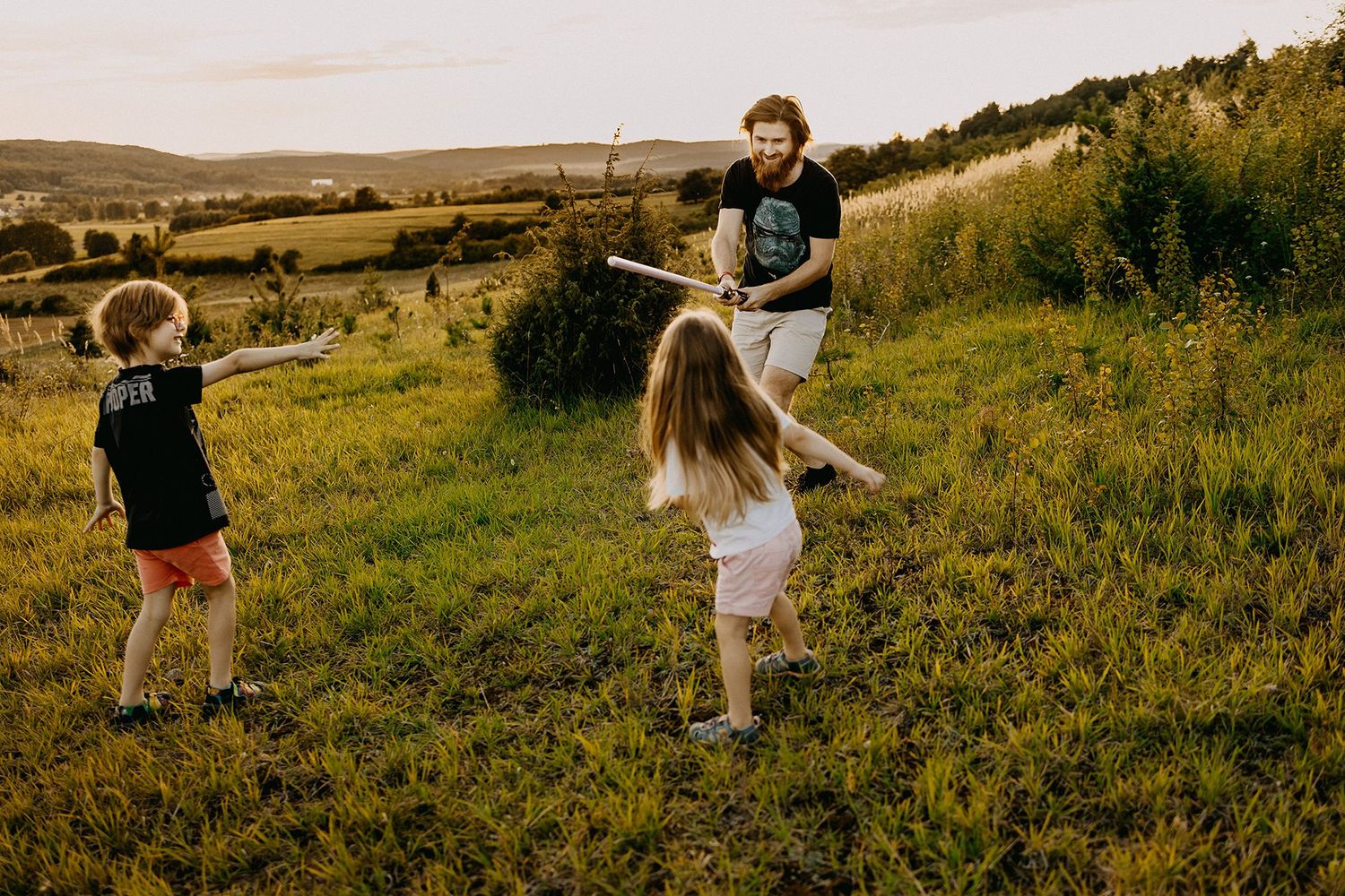 Father playfully sword fighting with two children on a green hillside at sunset in a scenic countryside.