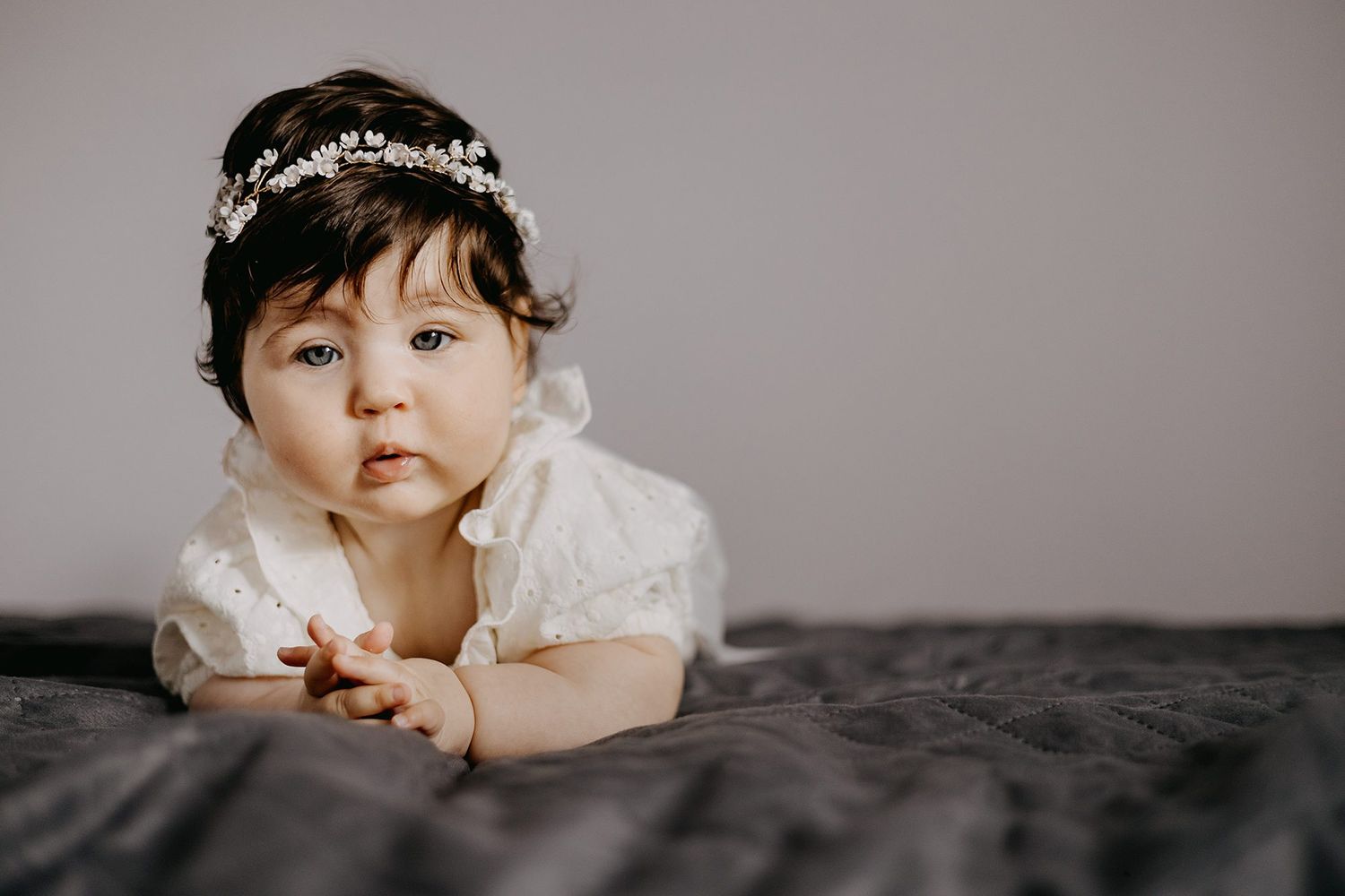 Adorable baby girl wearing white dress and floral crown lying on gray blanket looking at camera.