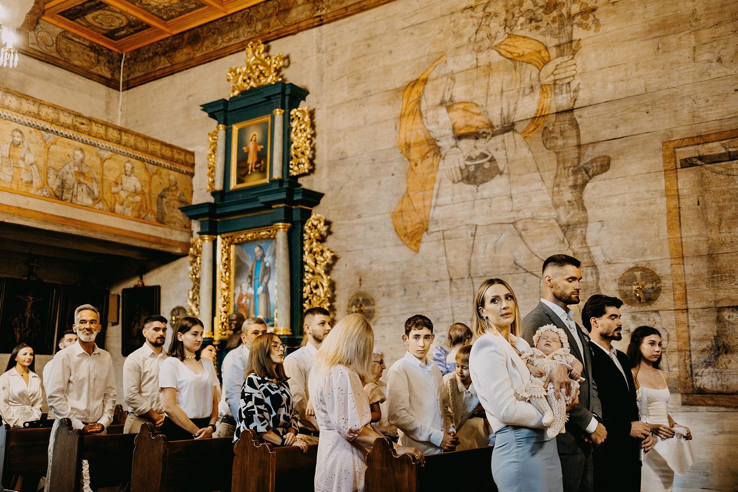 Family gathering in ornate church with golden altar and frescoed walls during baptism ceremony.