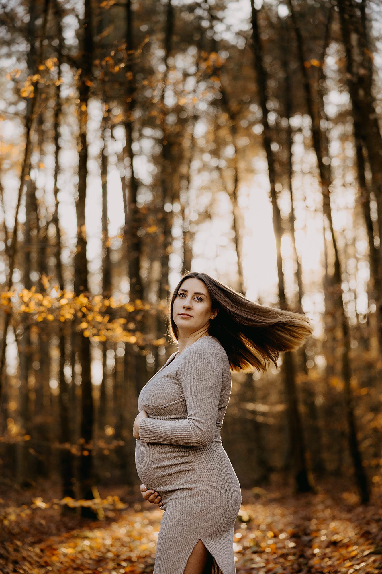 A pregnant woman in a grey dress poses gracefully in an autumn forest with golden foliage background.