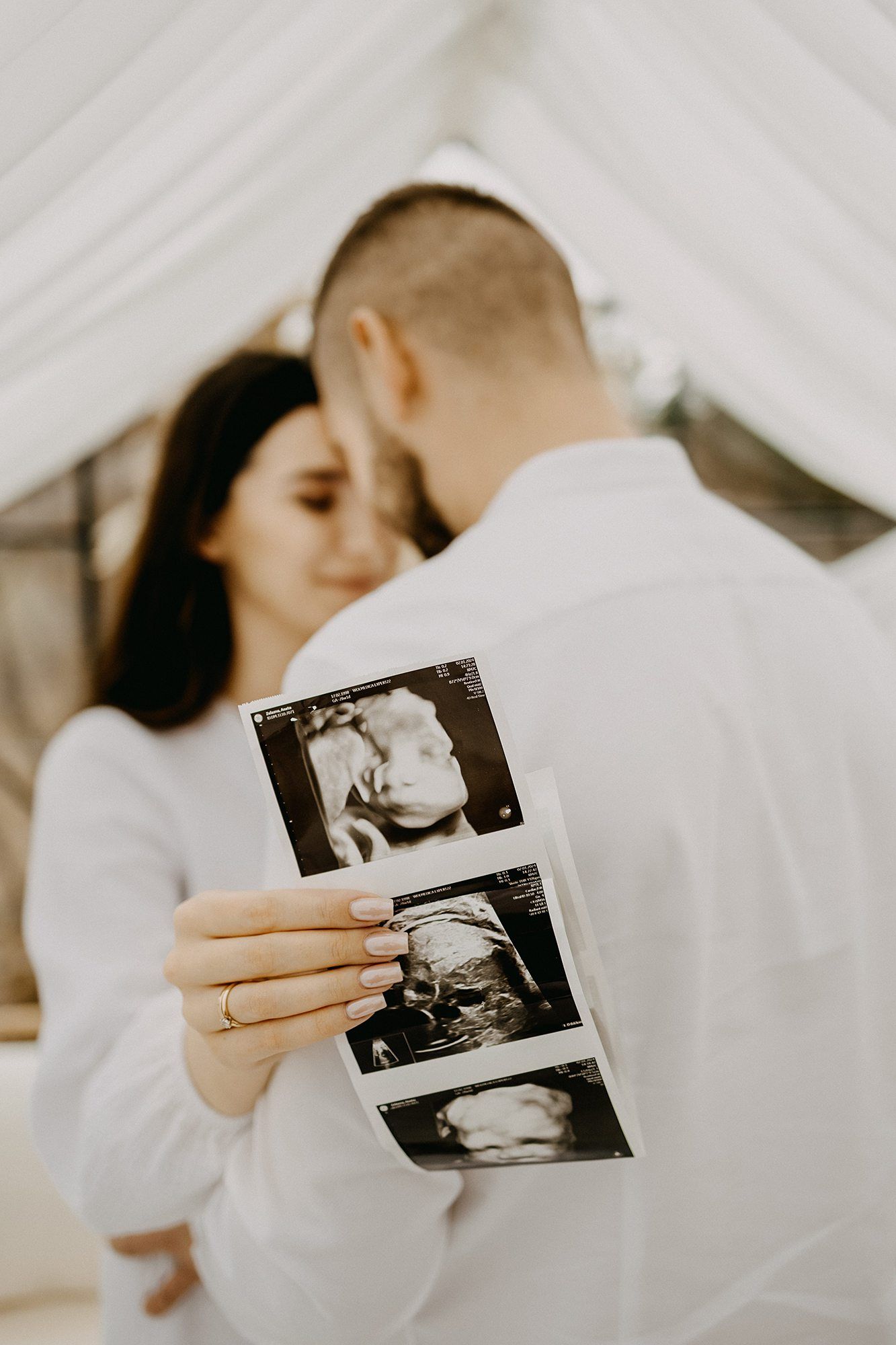 Pregnant couple holding ultrasound photos while embracing under white draped canopy.