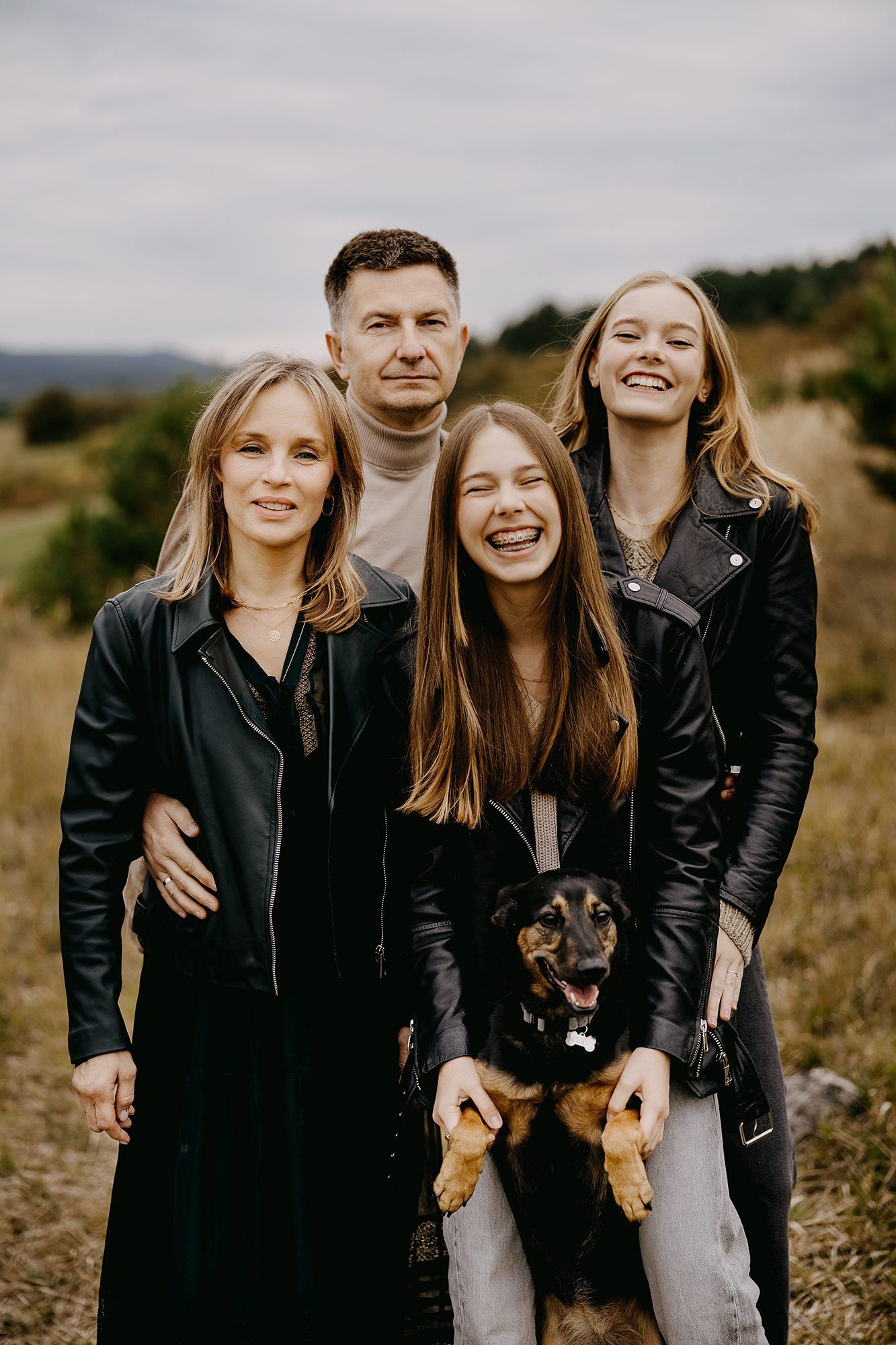 Happy family of four with a dog posing in an autumn field with rolling hills behind them.