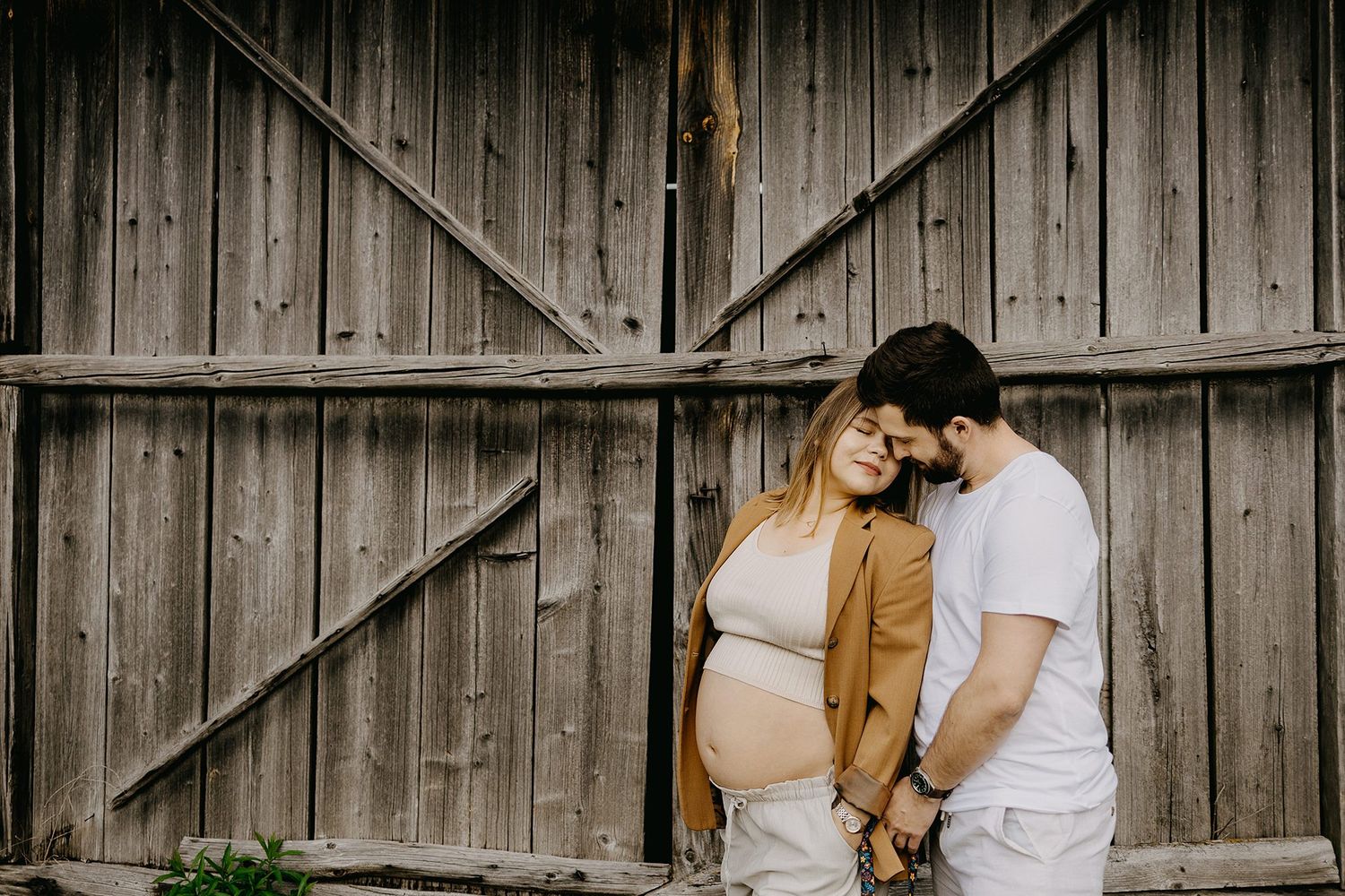 Pregnant couple leaning together holding hands against rustic wooden barn wall outdoors.