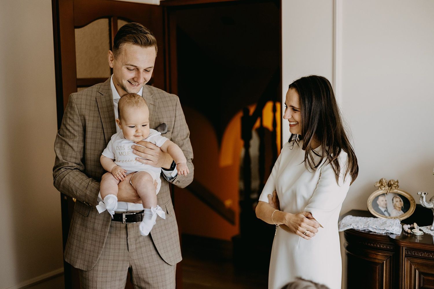 Man in plaid suit holding baby while woman in white dress smiles during christening celebration at home.