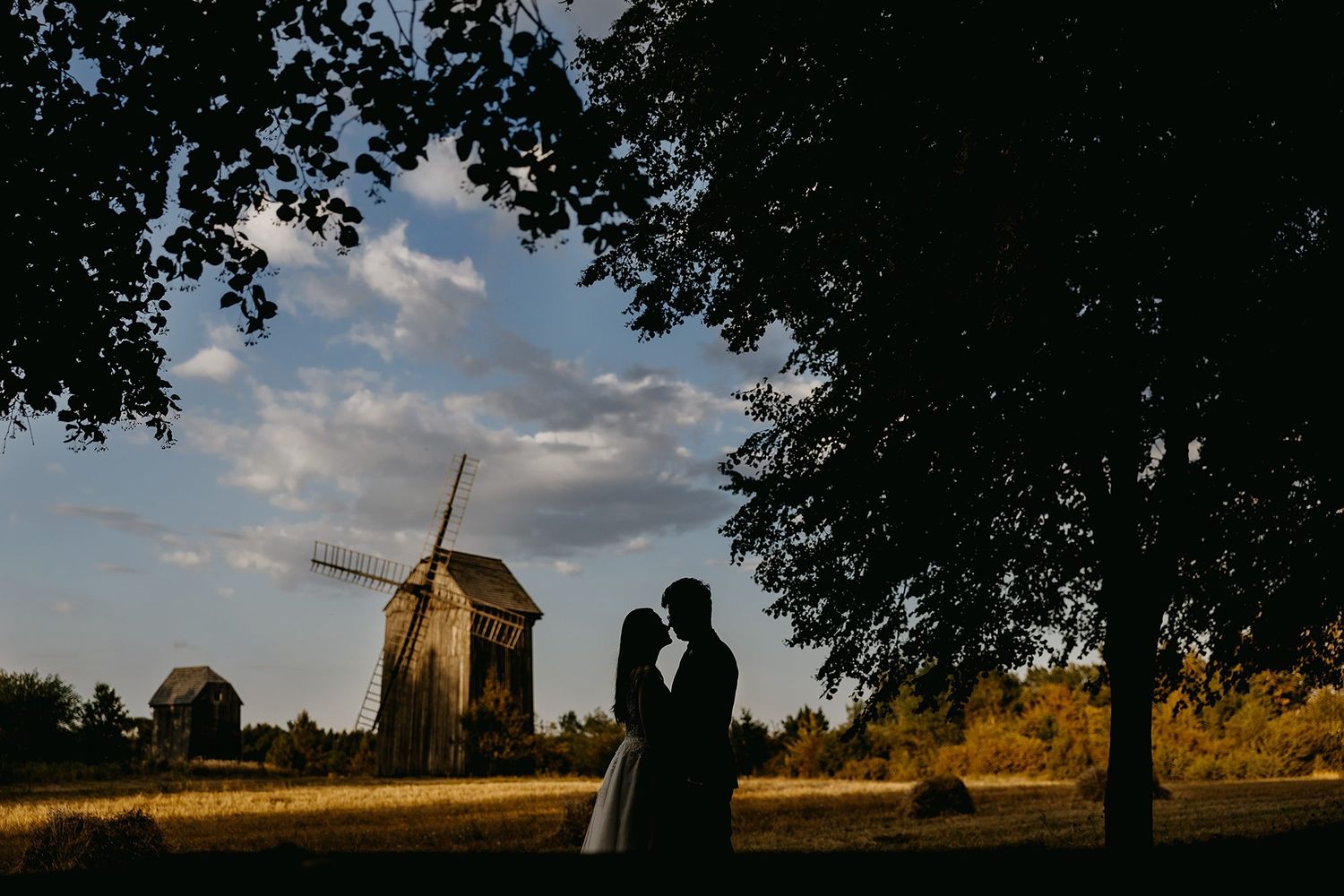 Romantic silhouette of bride and groom embracing near windmill at golden sunset under leafy trees.