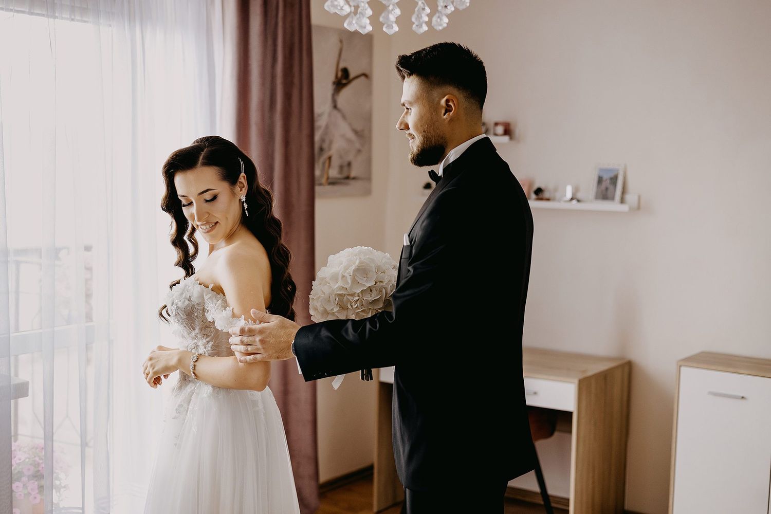 Groom in black tuxedo helping bride with wedding dress during pre-ceremony bridal preparations.