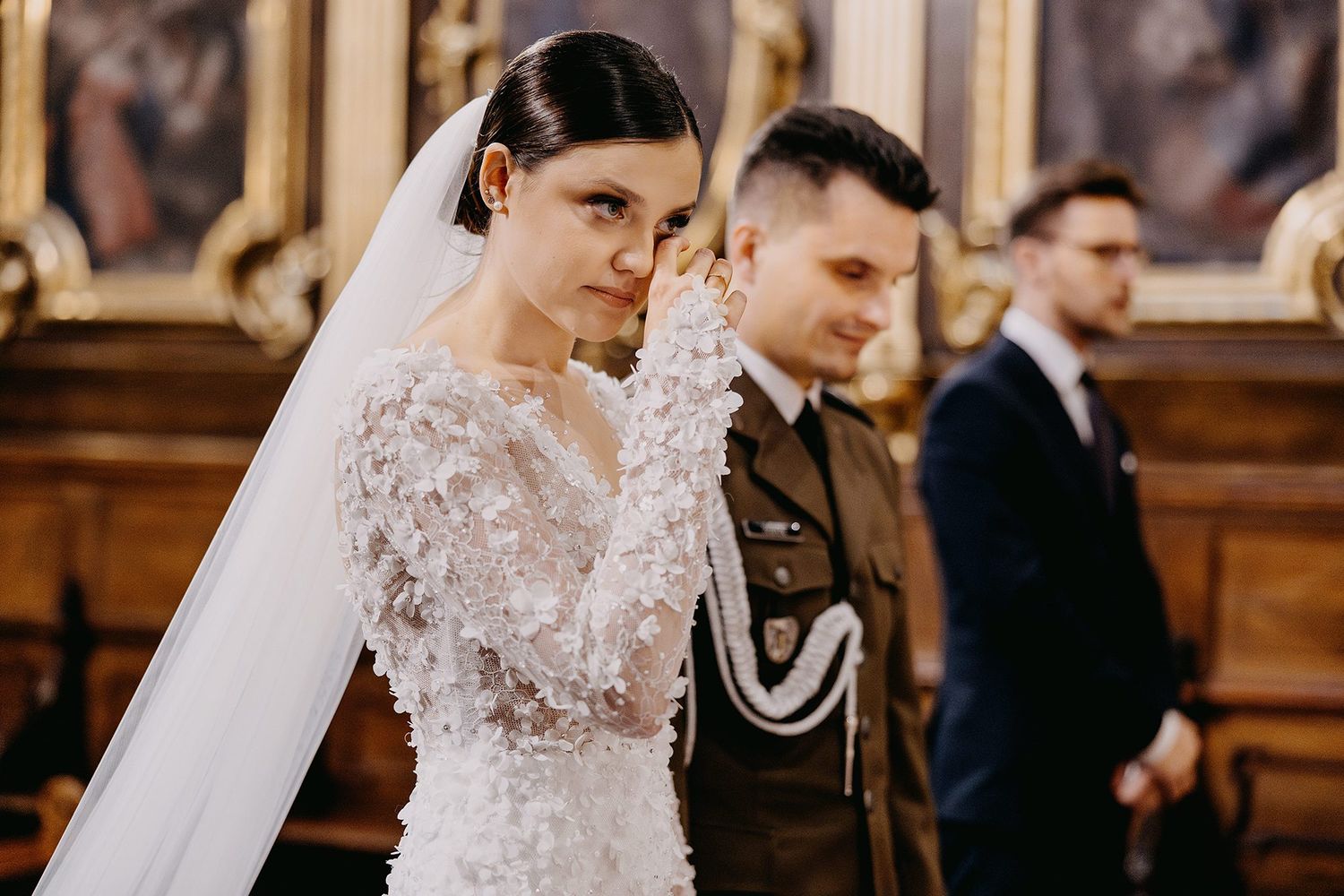 Emotional bride in lace wedding gown and veil wiping tears during church wedding ceremony.