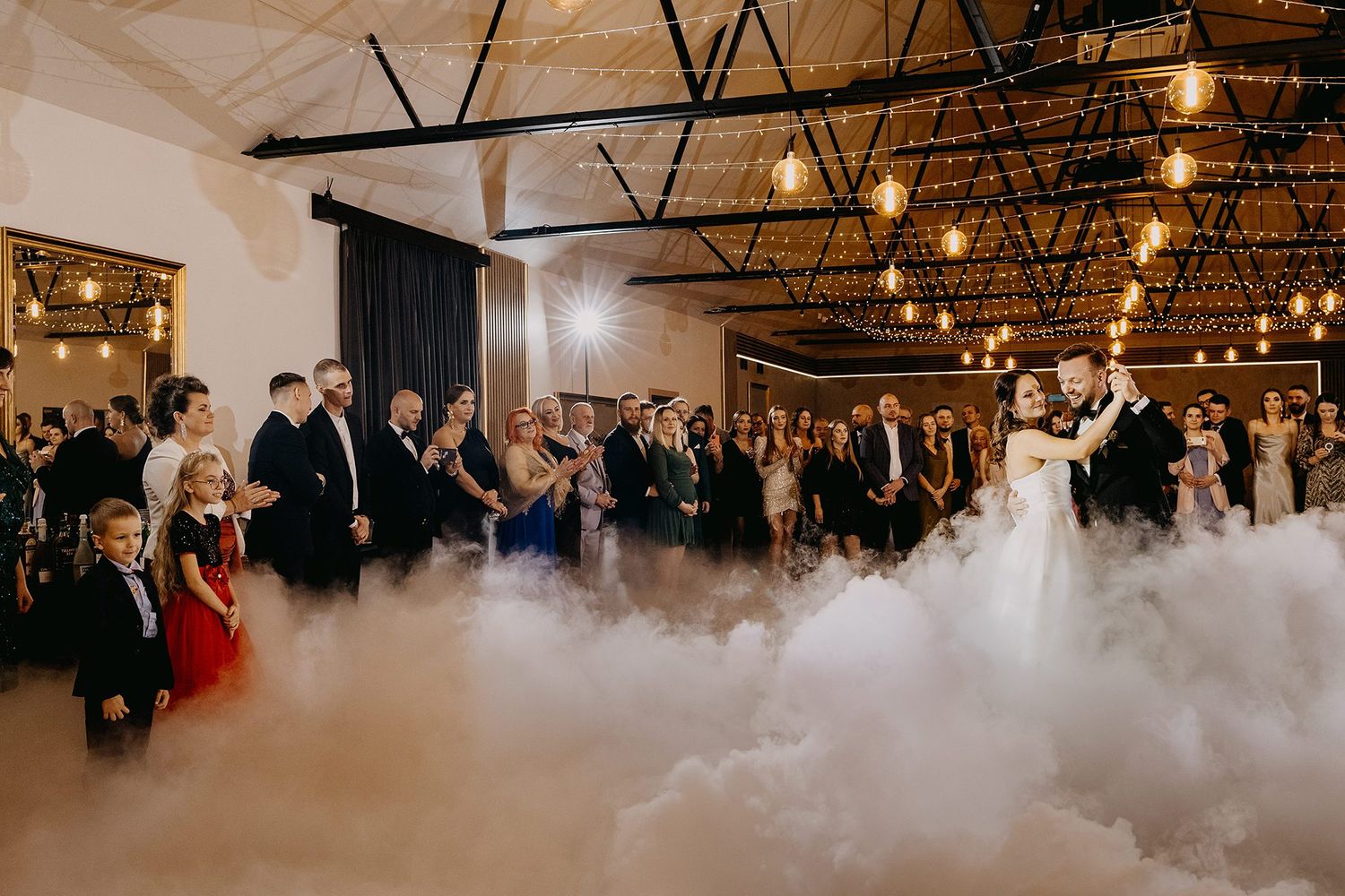Newlyweds share first dance surrounded by guests on a fog-filled dance floor under warm lights.