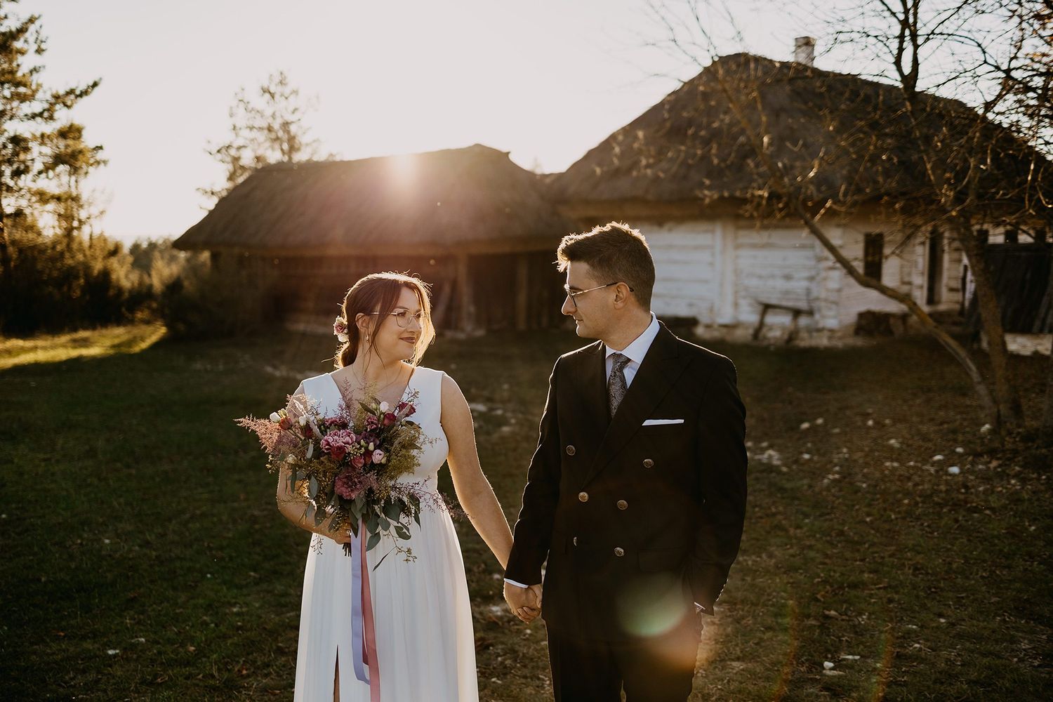 Bride and groom holding hands walking at golden sunset near rustic farmhouse, bride holding colorful bouquet.