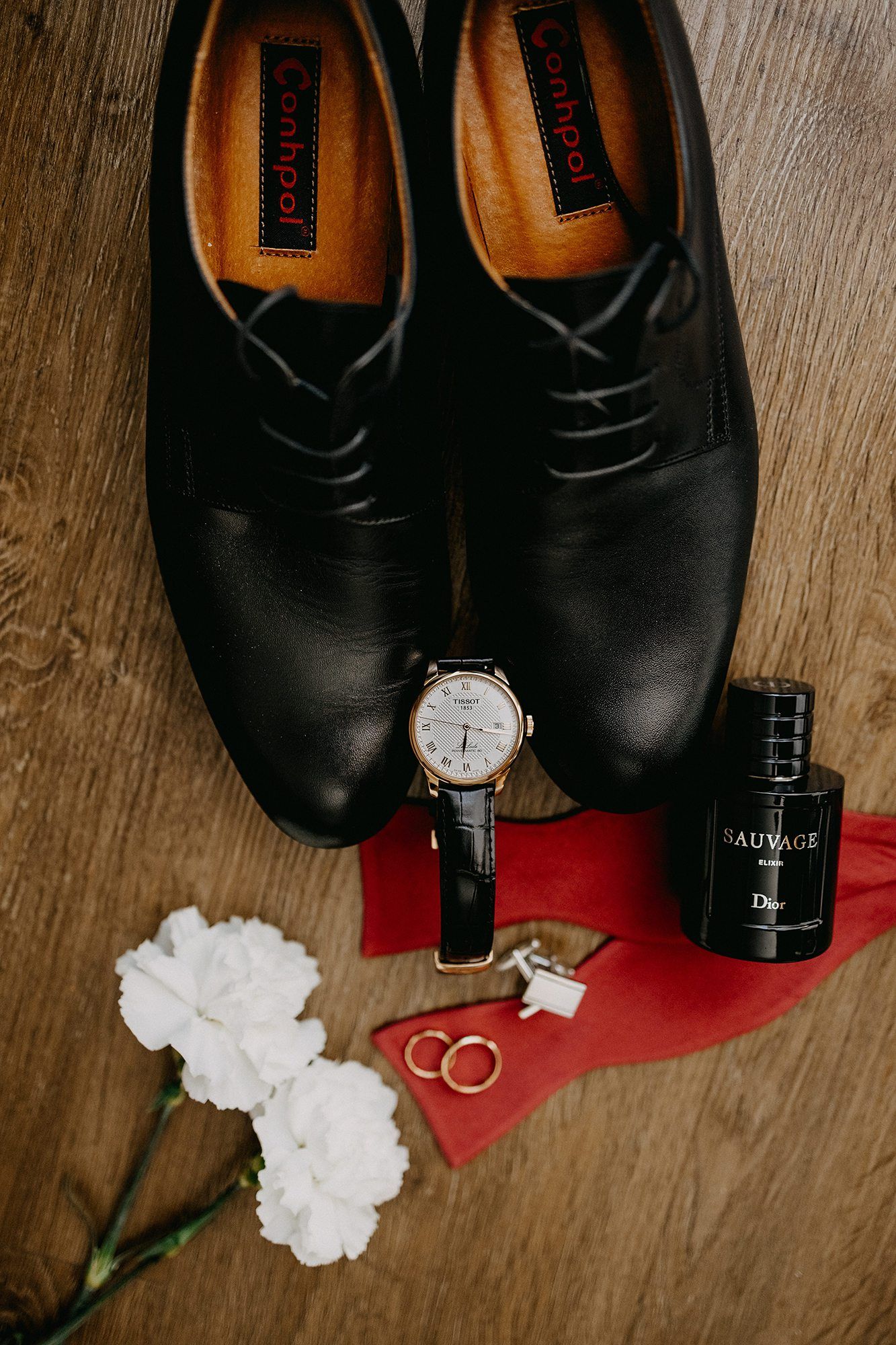 Elegant groom wedding accessories including black Oxford shoes, IWC watch, Dior Sauvage cologne, gold rings, and white carnations.