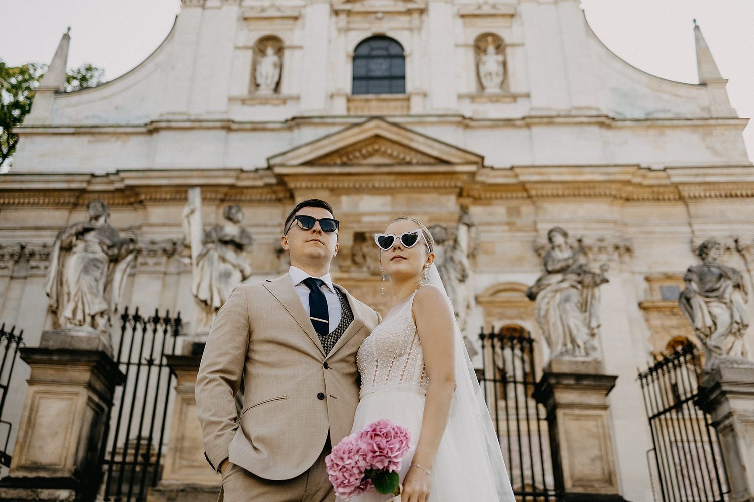 Stylish bride and groom wear sunglasses posing before a baroque church with pink bouquet.
