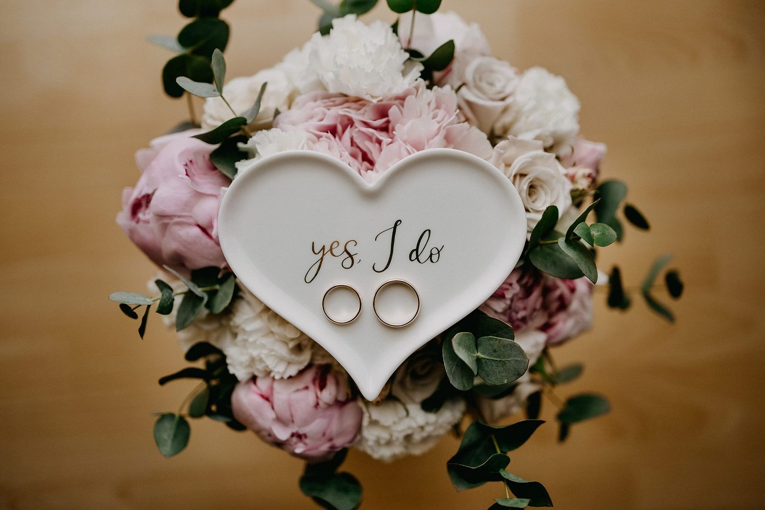 Two gold wedding rings on a heart dish with yes I do text atop a floral bouquet.