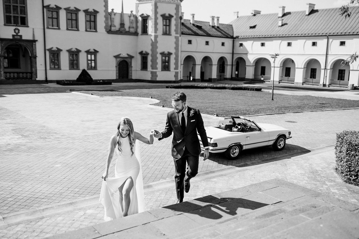 Black and white aerial view of bride and groom walking hand in hand near classic convertible.
