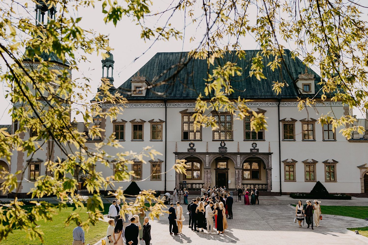 Wedding guests gather outside a historic European manor building surrounded by spring foliage.