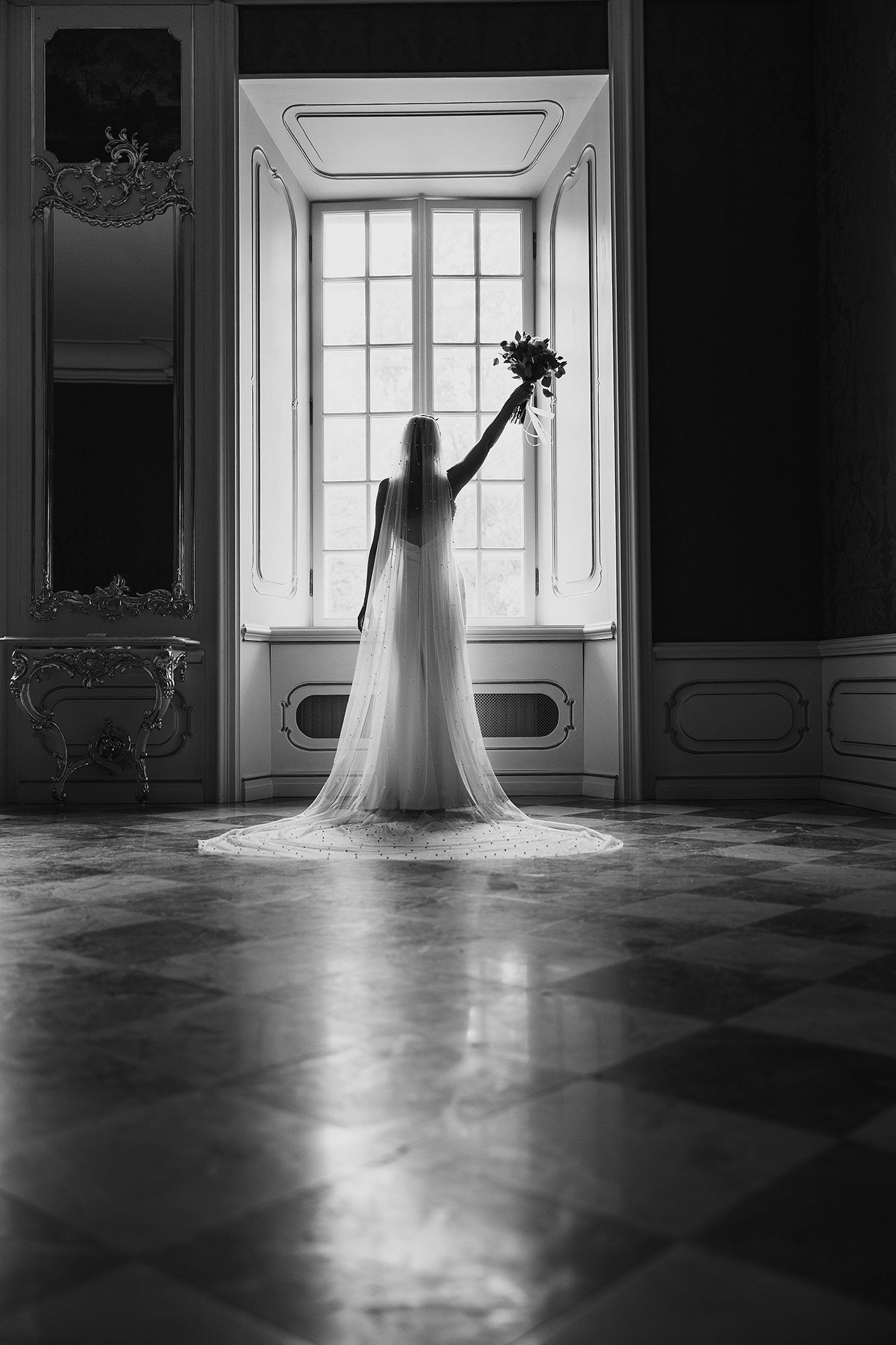 Bride in wedding gown holding bouquet aloft before grand window in elegant black and white photo.
