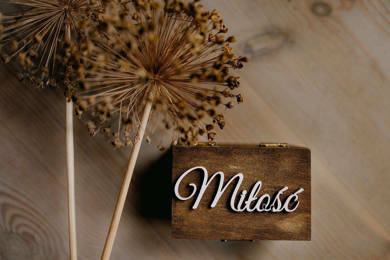 Rustic wooden ring box engraved with Milosc beside dried allium flowers on wood surface.