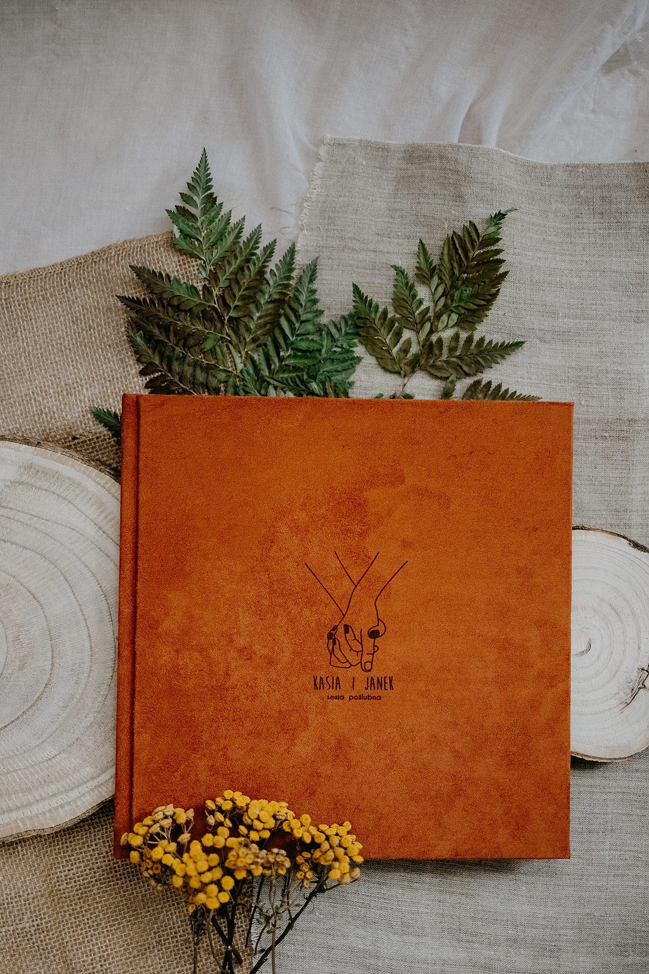 Orange velvet wedding photo album with couple illustration on linen beside fern and yellow flowers.