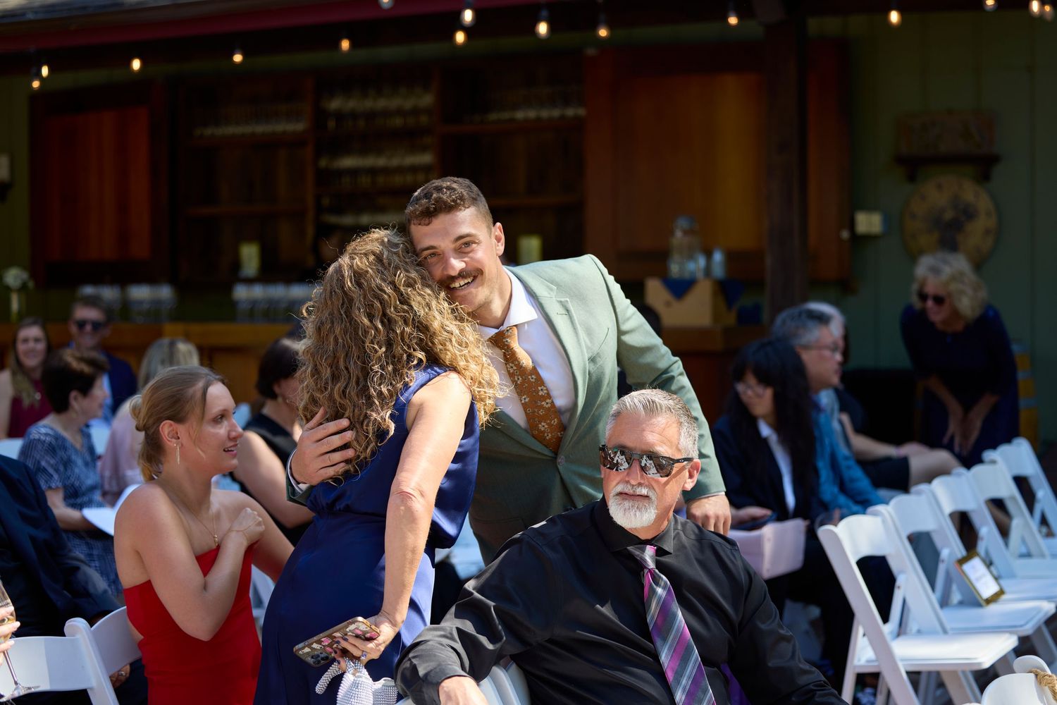 Wedding guest in mint green suit greeting seated guests at an outdoor ceremony with white chairs.