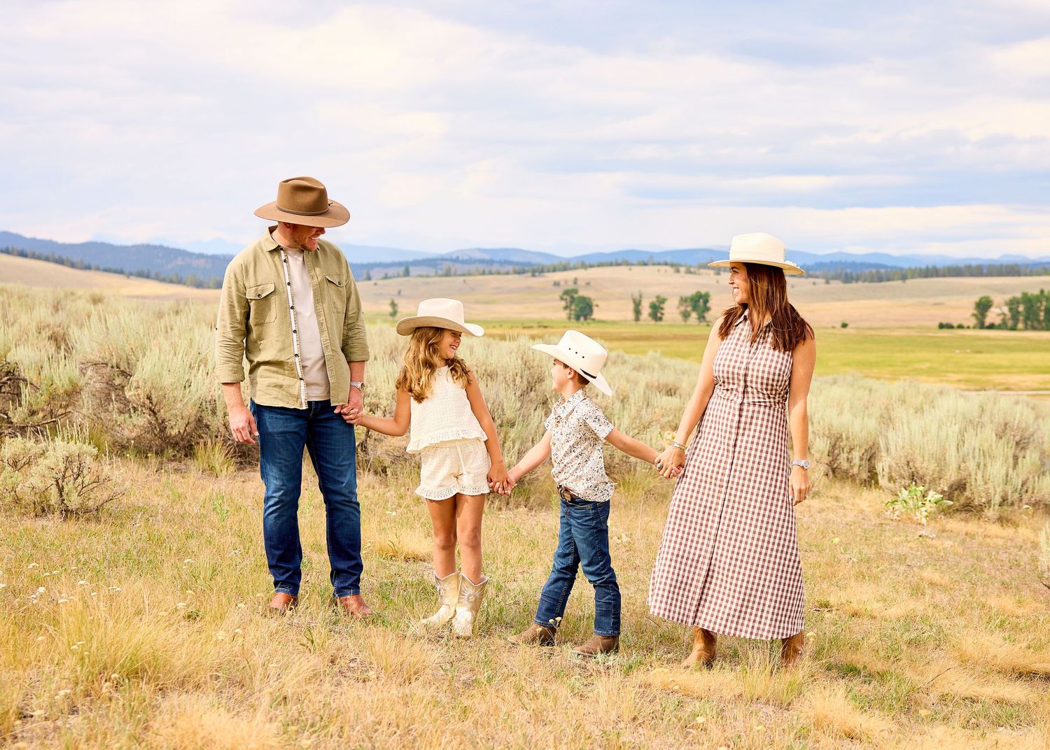 Family of four in western hats holding hands walking through a sagebrush meadow under cloudy skies.