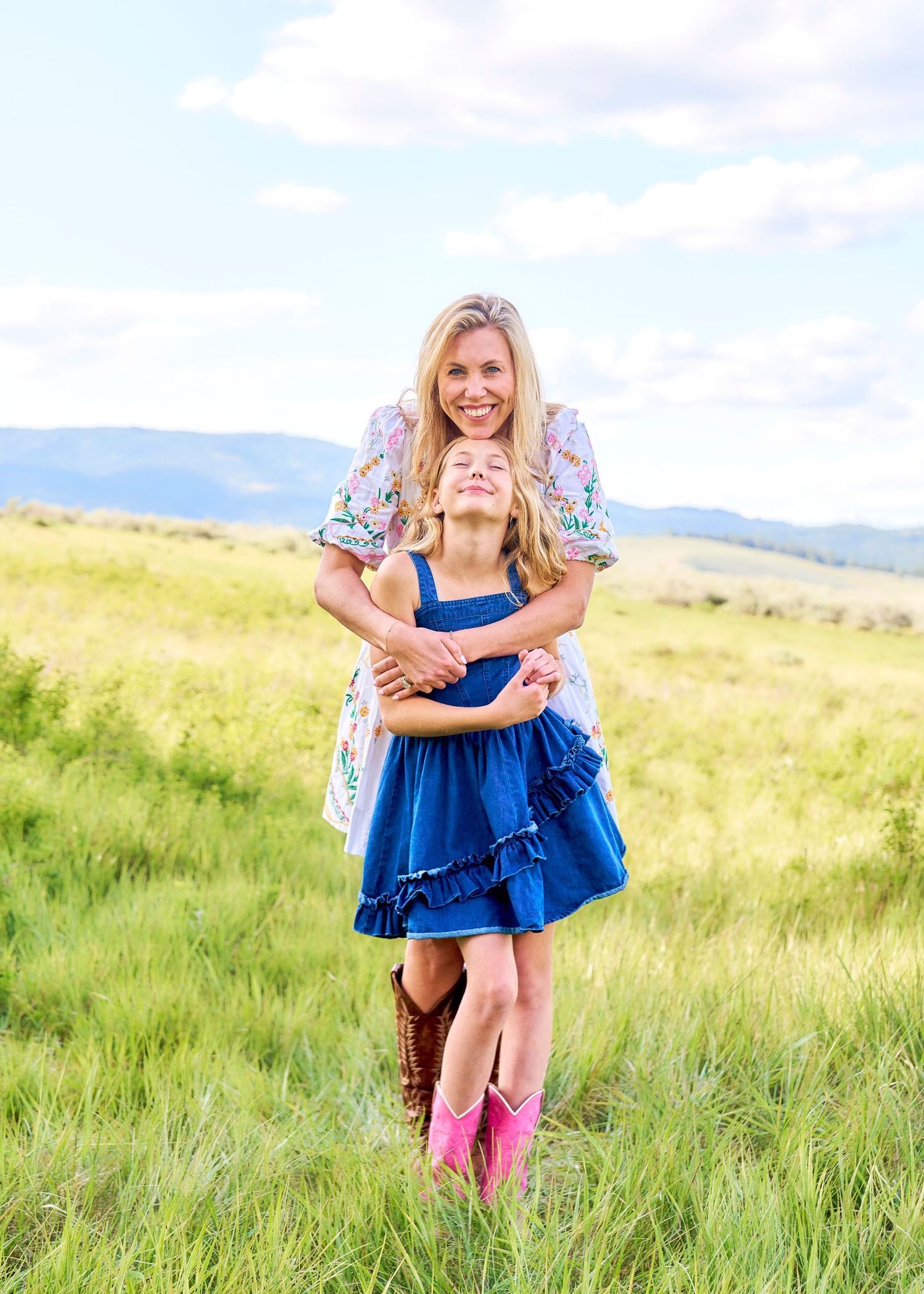 Blonde woman hugging young girl in blue dress and pink boots in green mountain field.