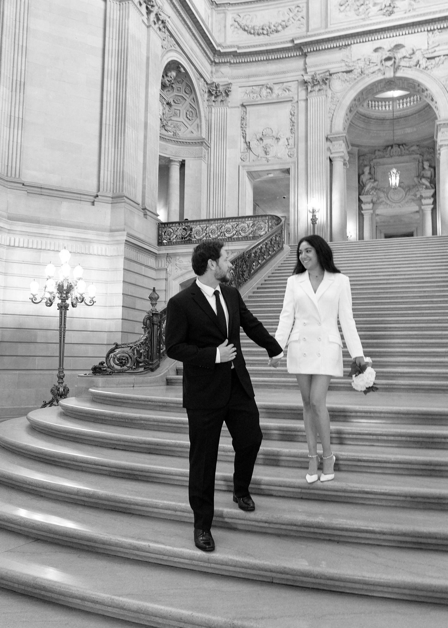 Black and white wedding photo of couple holding hands walking down grand staircase in San Francisco City Hall.