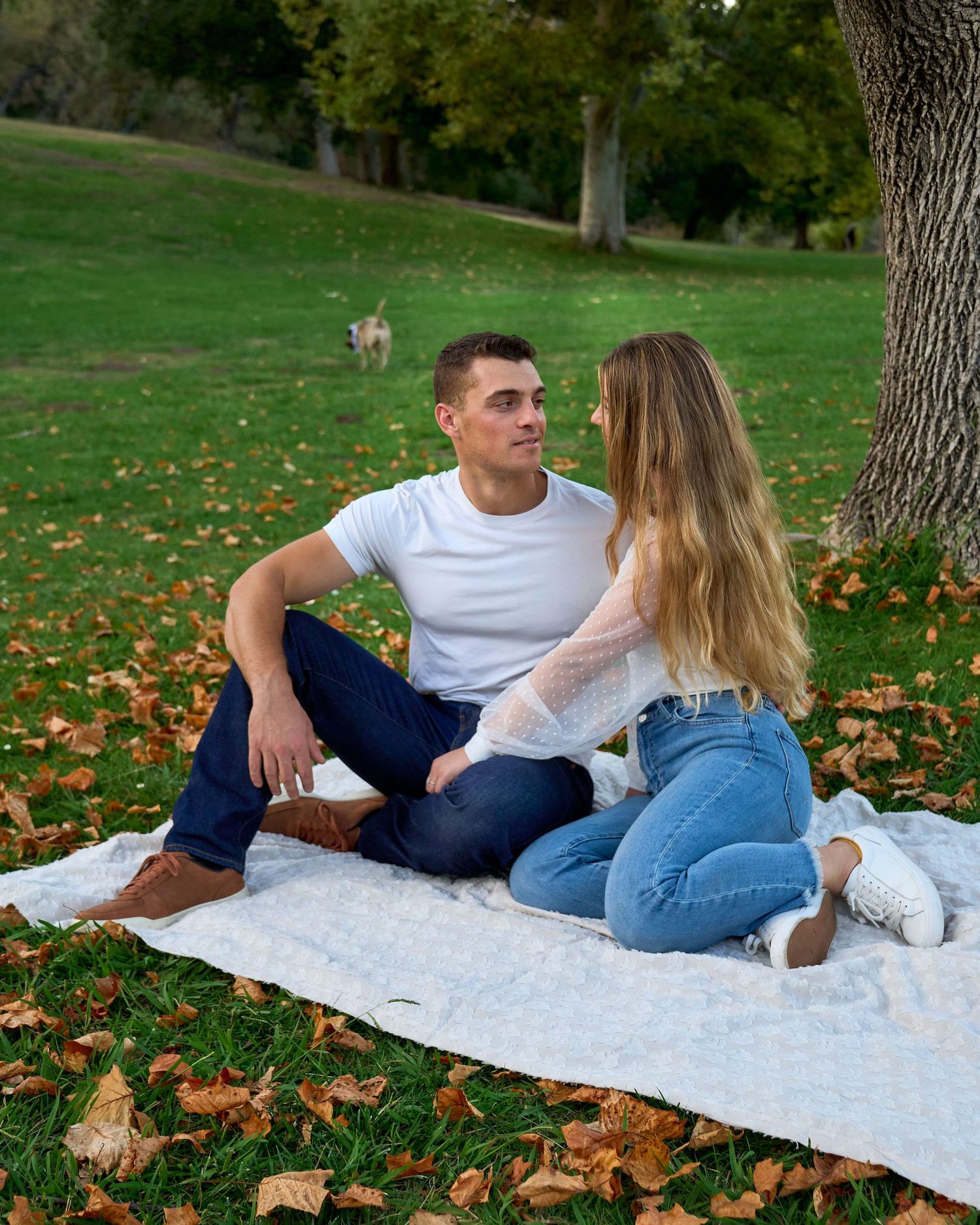Young couple sitting on blanket in autumn park surrounded by fallen leaves with dog in background.