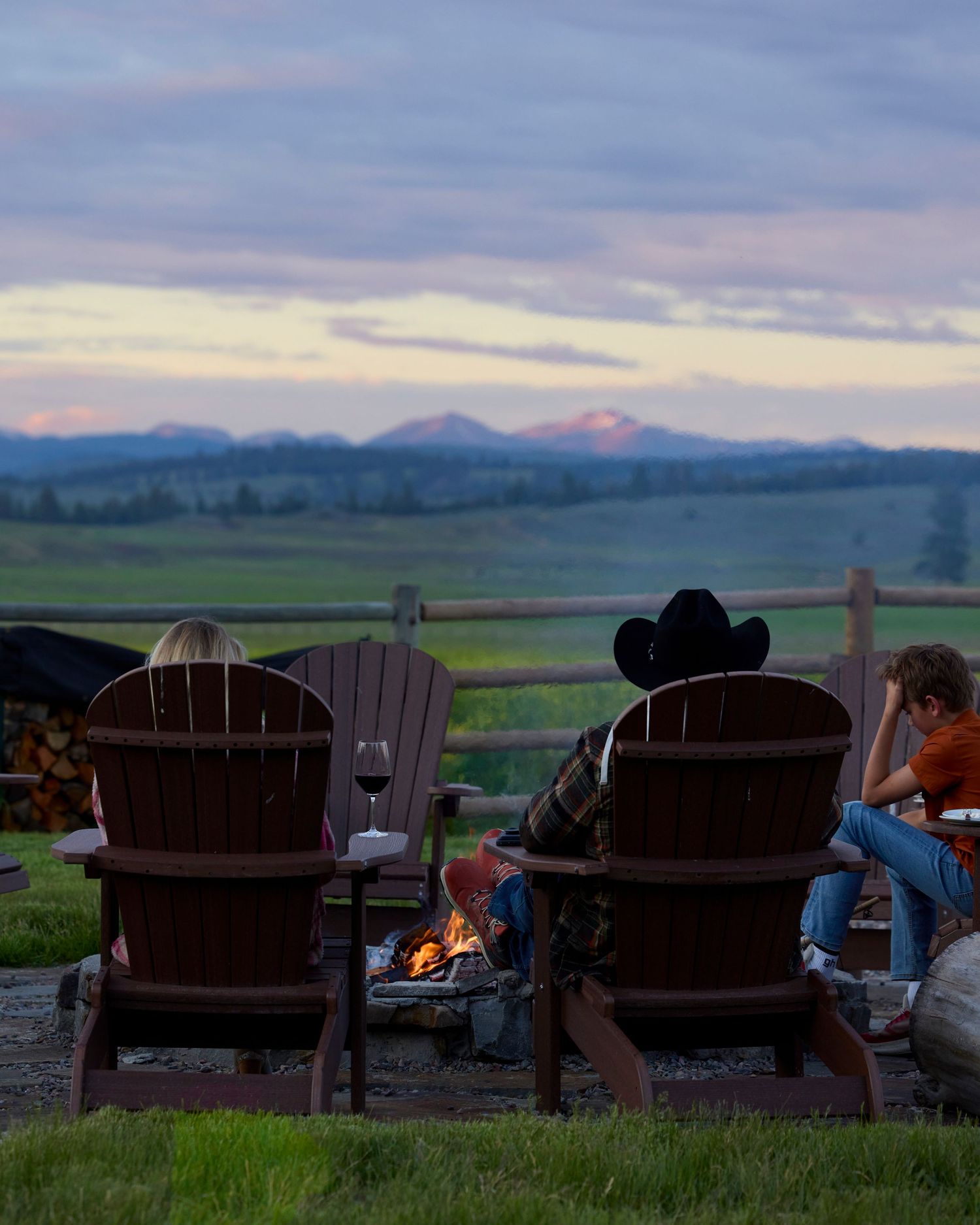 Two people in Adirondack chairs sit by a campfire overlooking a mountain valley at sunset.