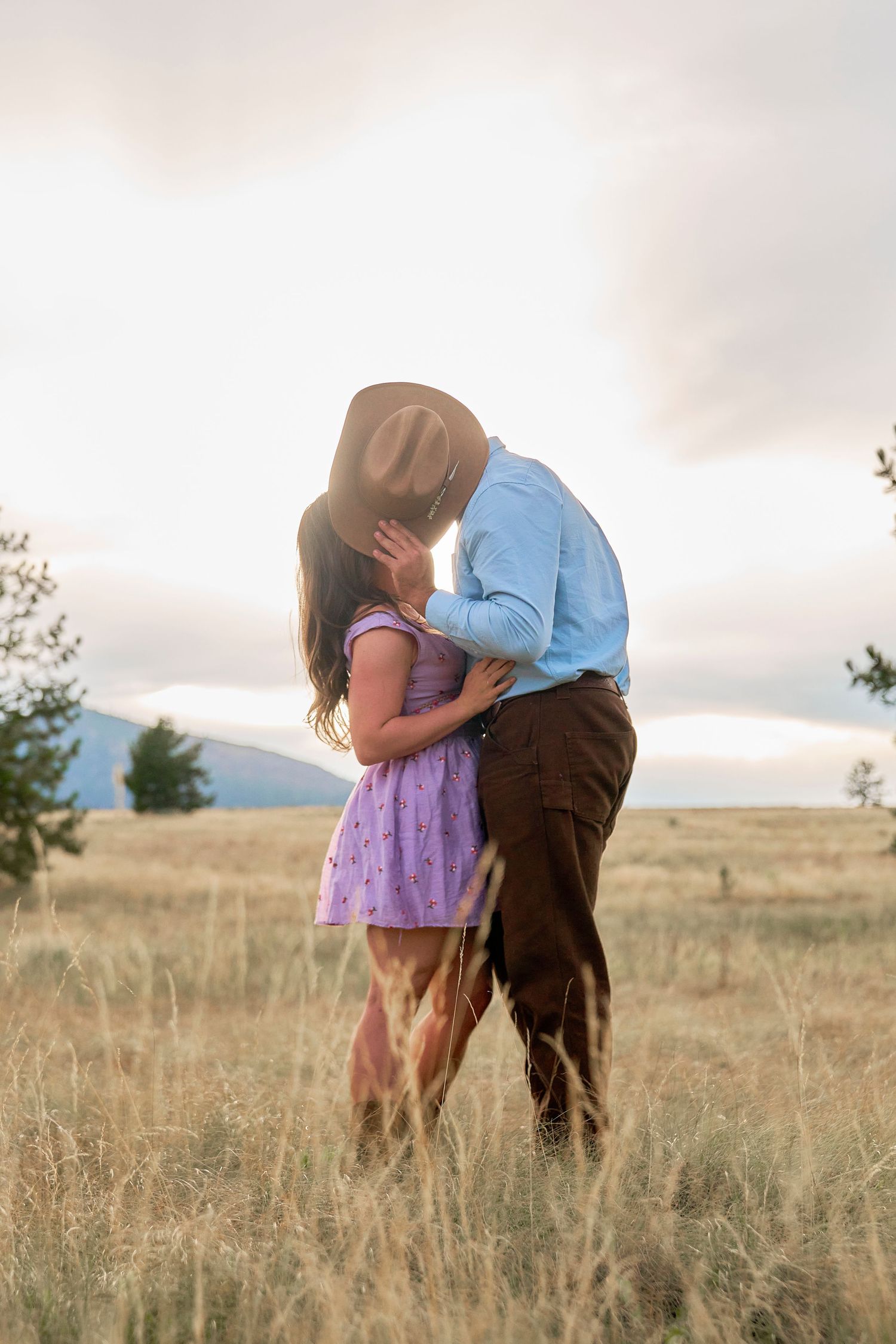 Couple sharing a kiss in a sunlit golden meadow, man in cowboy hat and blue shirt holding woman in purple dress.