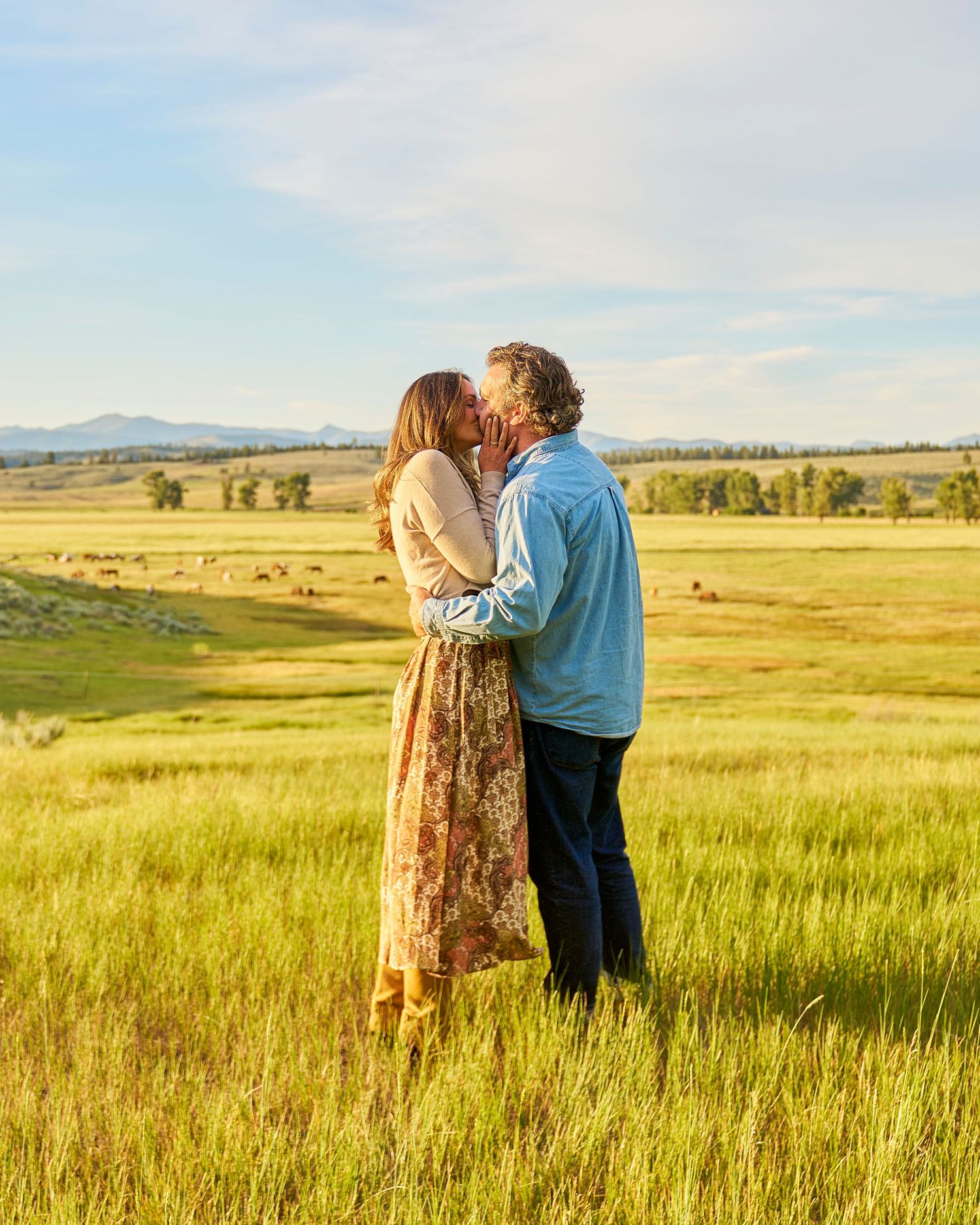 Romantic couple sharing a kiss in a golden sunlit meadow with mountains and grazing cattle in the background.