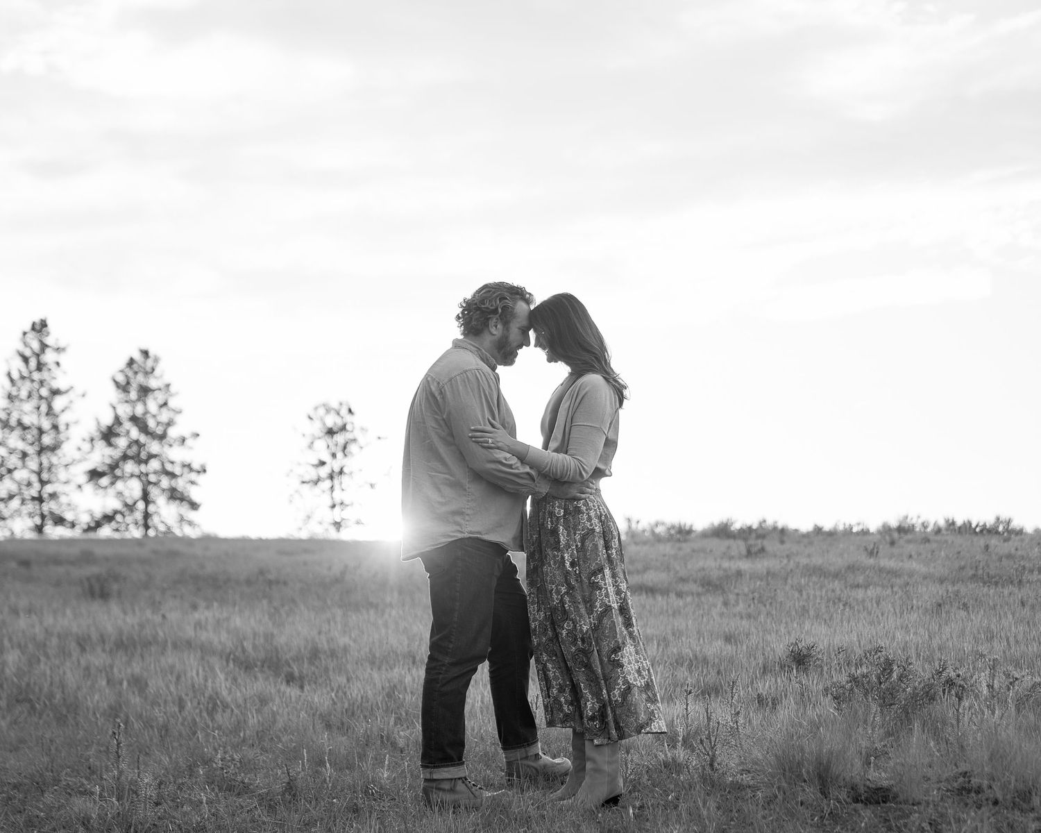 Black and white photo of a romantic couple standing close together in a field with pine trees at sunset.