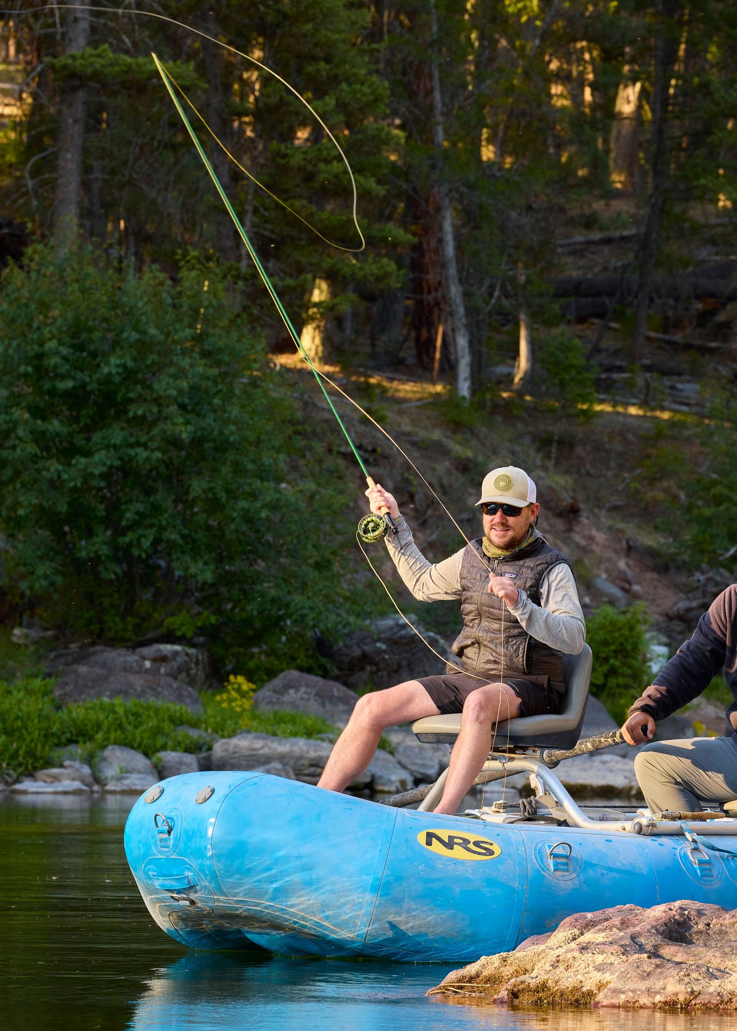 A smiling man fly fishing from a blue NRS raft on a scenic forested river.