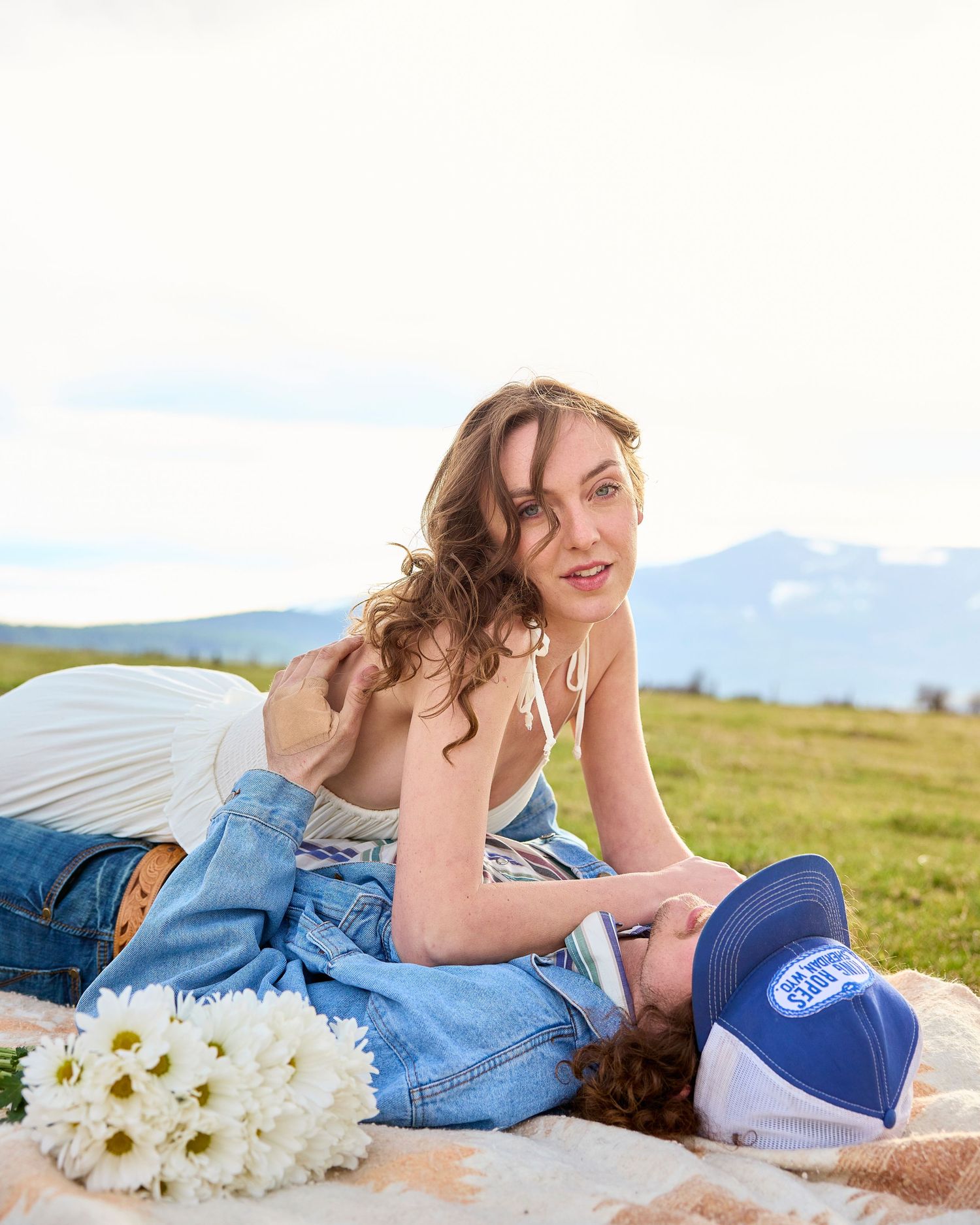 Bride in white dress posing over groom lying on blanket in green field with mountain scenery and daisies.