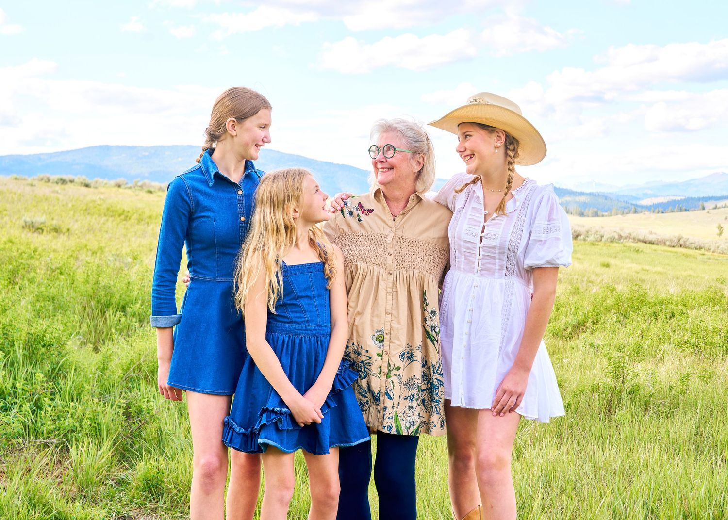 Three generations of women smiling together in a lush green field under cloudy skies.