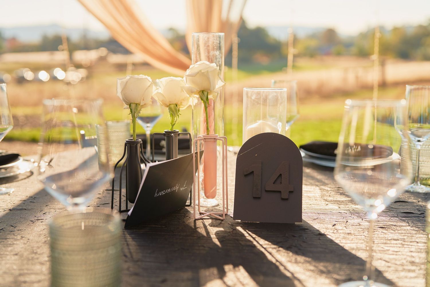 Elegant outdoor wedding reception table setting with white roses in test tubes, table number 14, and glassware in warm sunlight.
