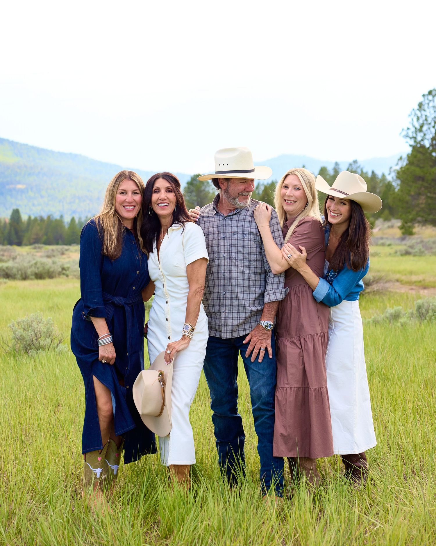 Five adults laughing and embracing in a green meadow with mountain and pine tree backdrop.