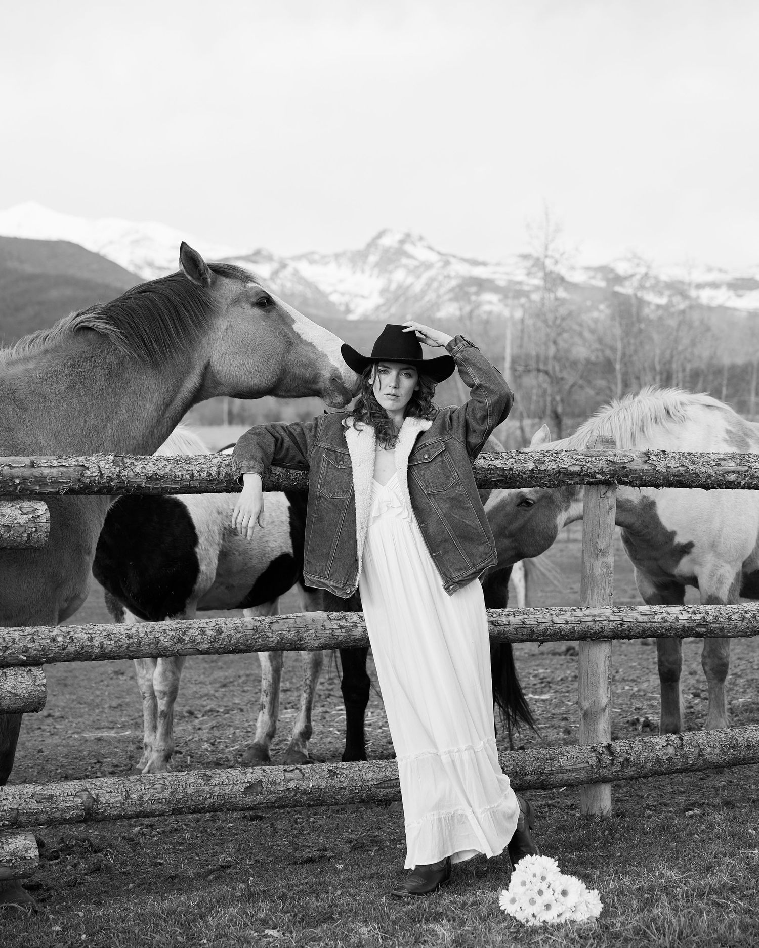 Bride in black cowboy hat and denim jacket poses with horses at ranch fence in black and white.