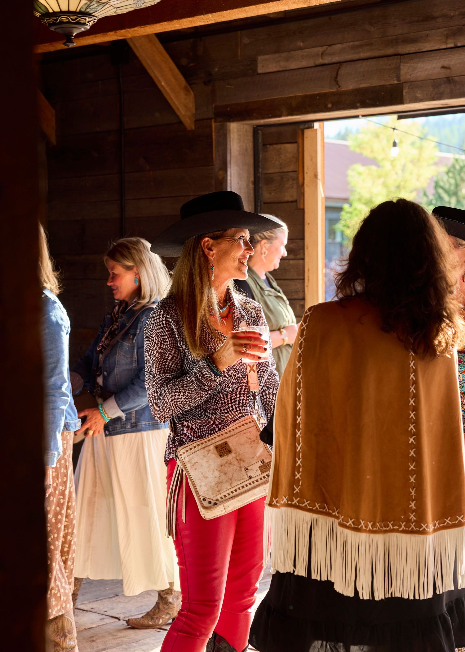 Stylish women socializing at a rustic western venue, one wearing a black hat and holding a drink.