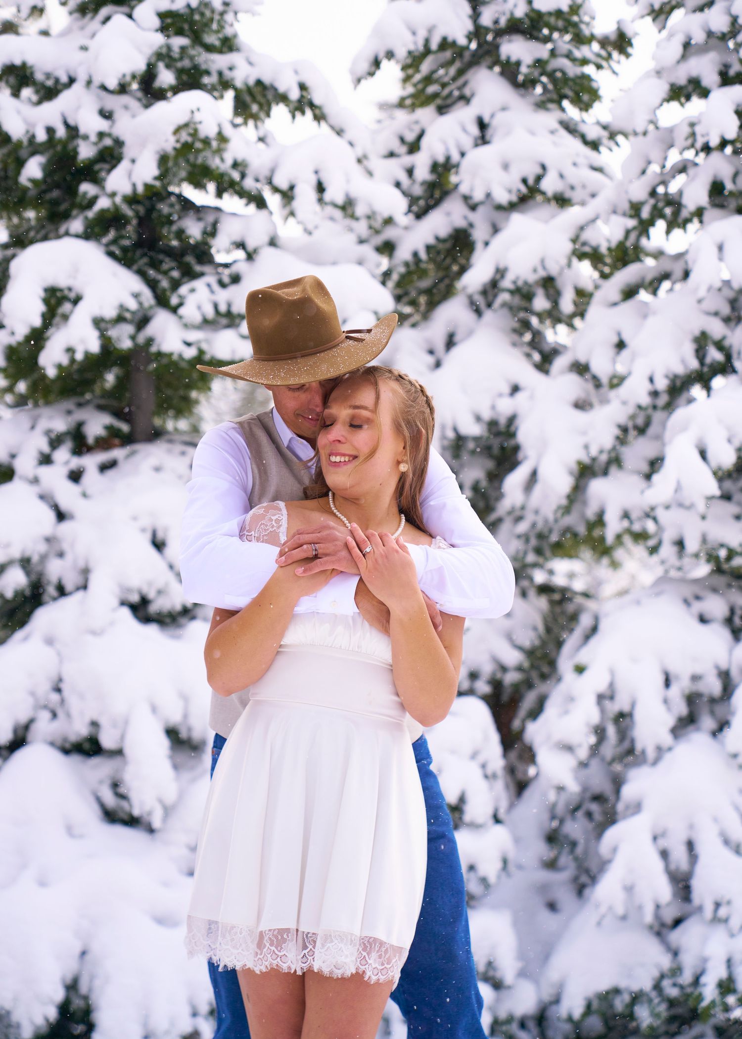 Groom in cowboy hat embraces bride in white mini dress before snowy pine trees in winter wedding photo.