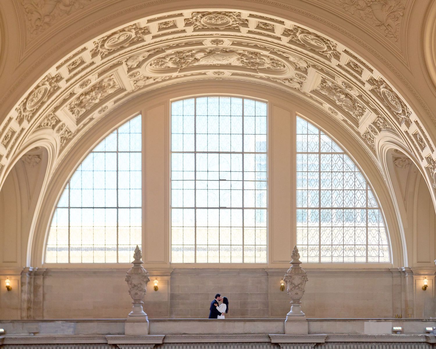 Wedding couple shares intimate moment on balcony beneath grand ornate arched window in elegant venue.