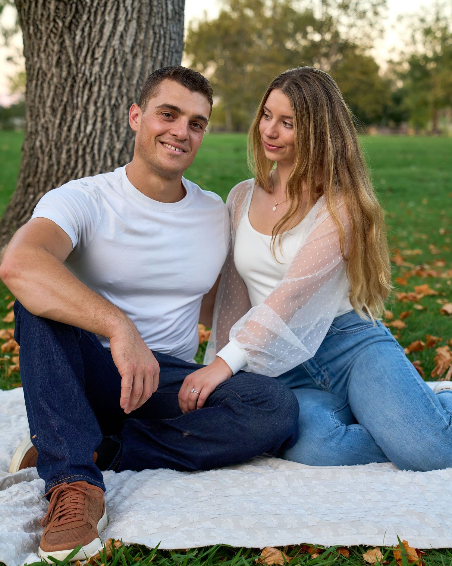Couple sitting together on white blanket under tree surrounded by fallen autumn leaves in park.