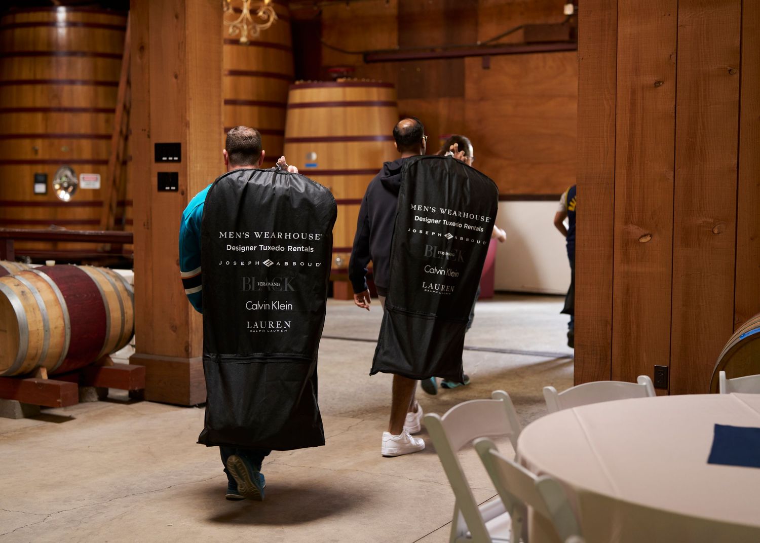 Two groomsmen in Men's Wearhouse garment bags walking through a winery barrel room before the wedding.