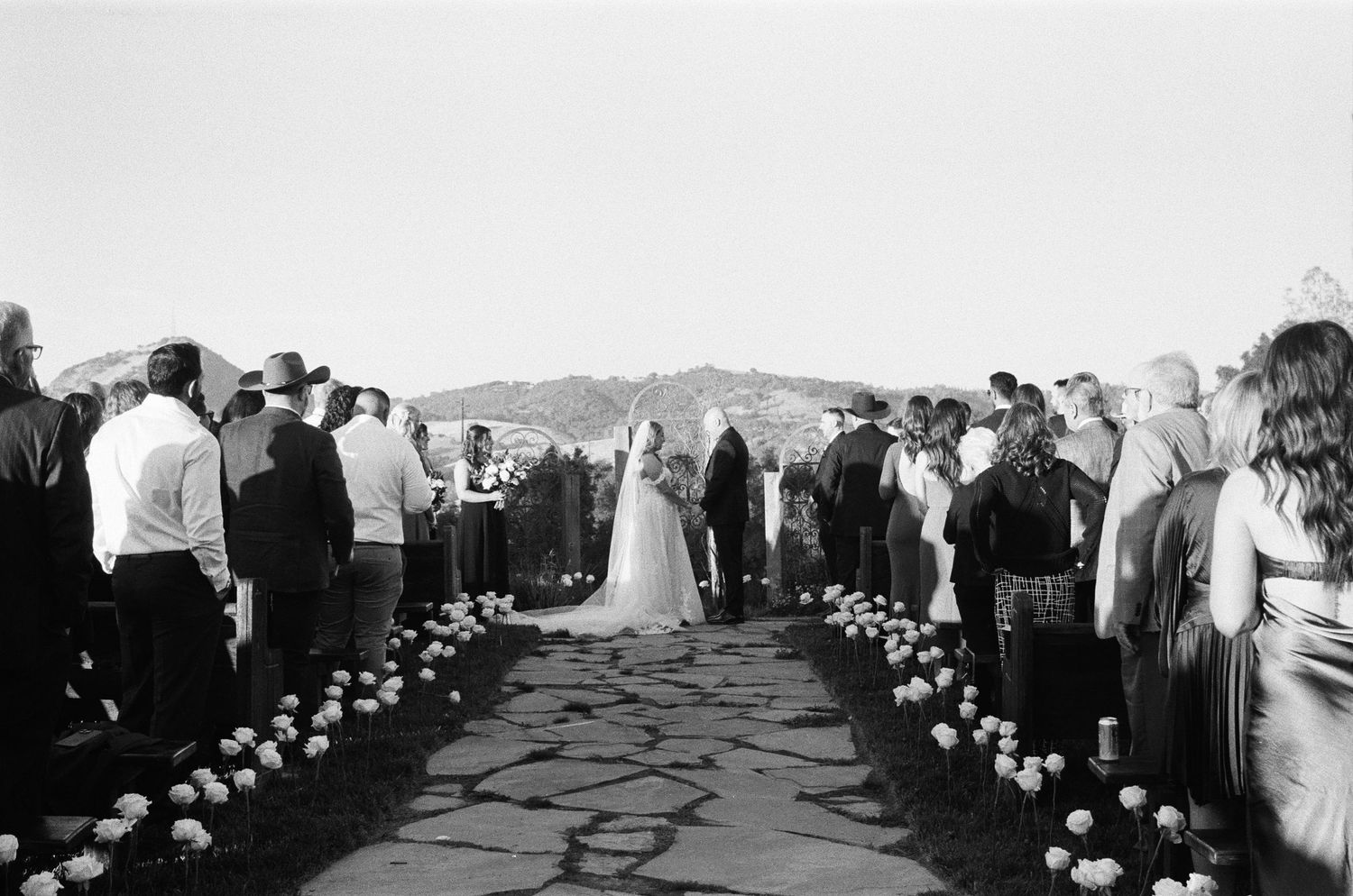 Black and white aerial view of outdoor wedding ceremony with guests lining a stone aisle with white roses.