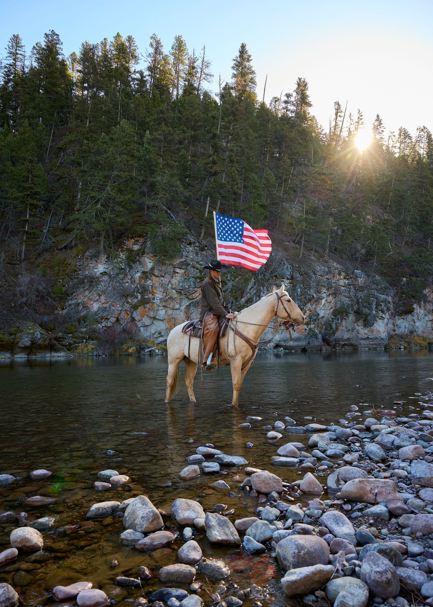Cowboy on white horse holds American flag while crossing a rocky river at golden sunset.