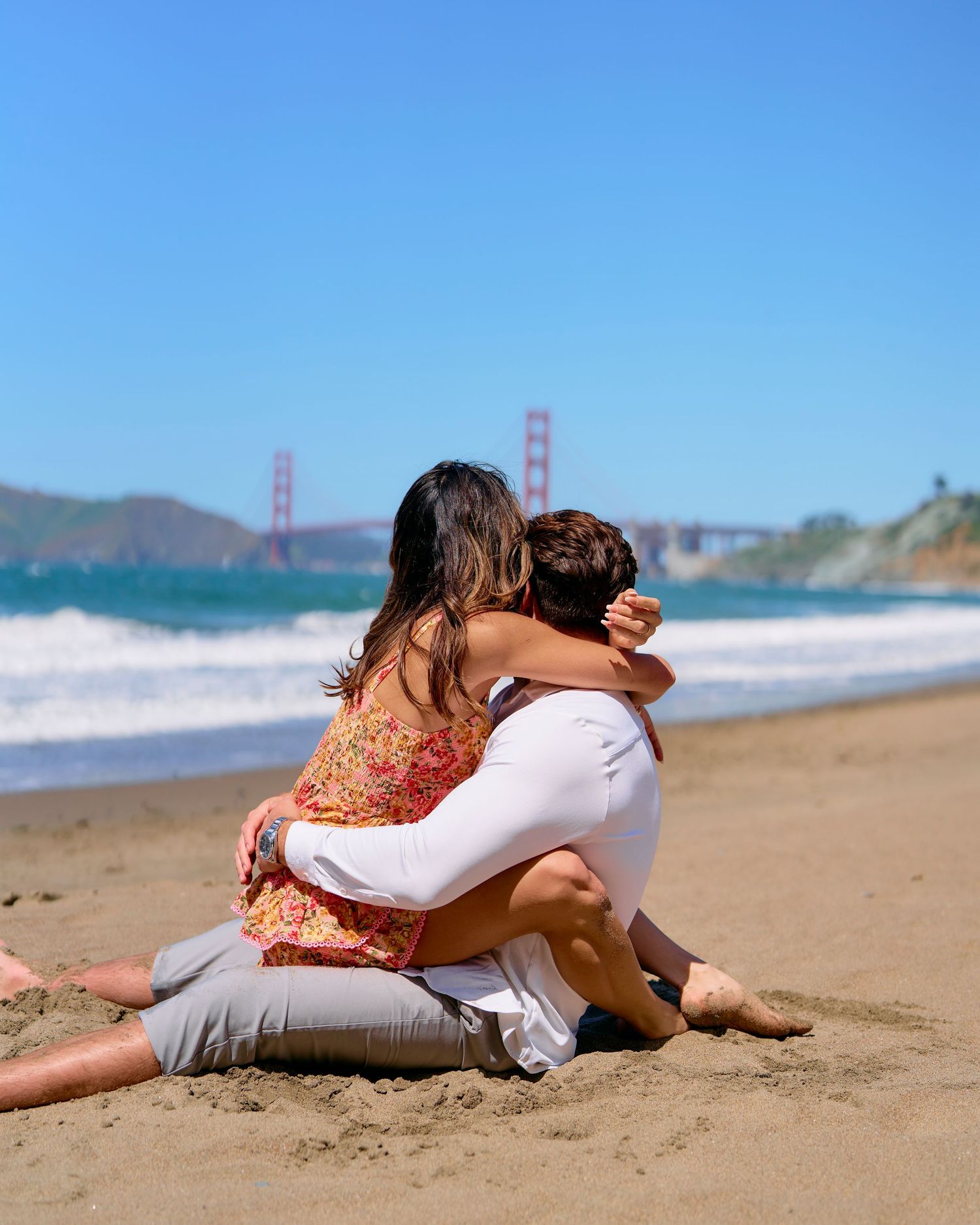 Couple sitting together on Baker Beach sand with Golden Gate Bridge visible in background.