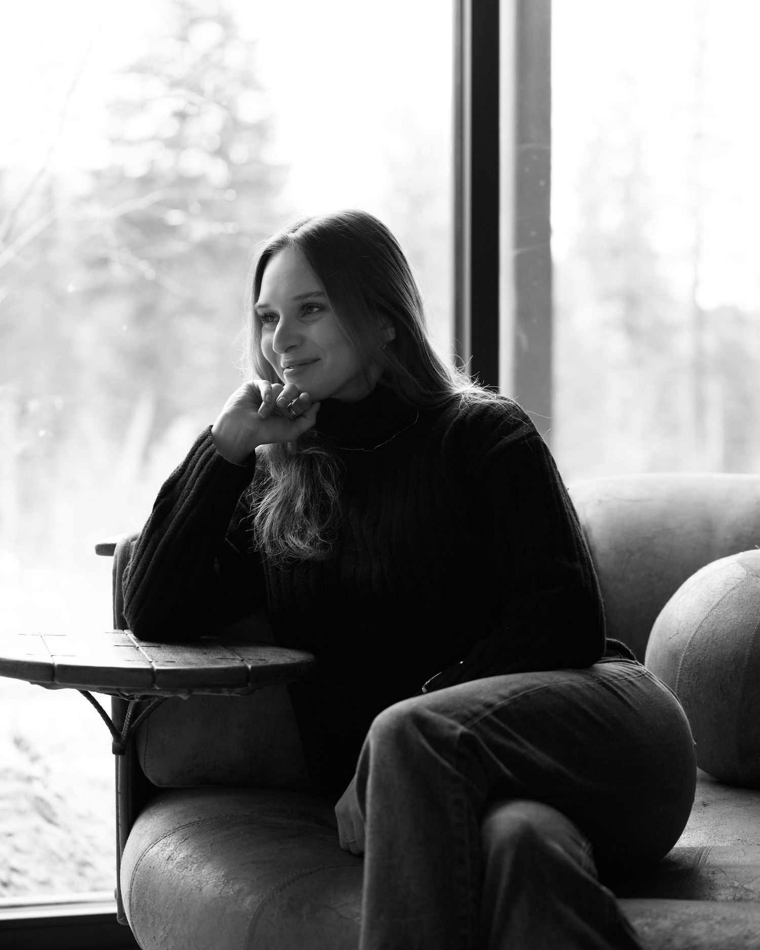 Black and white portrait of woman with long hair relaxing in armchair by large window.