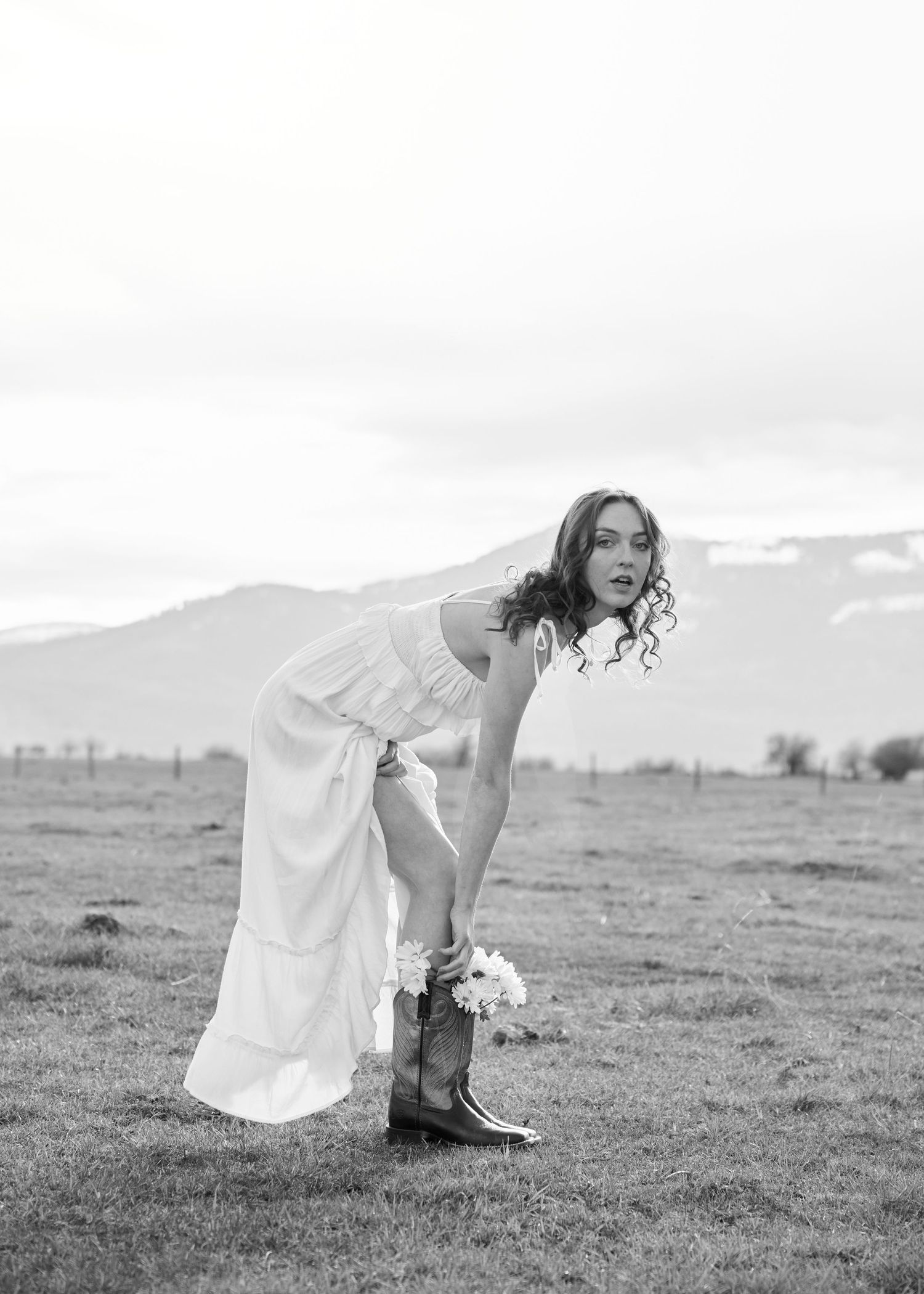 Bride in white dress and cowboy boots bends down holding flowers in open field, black and white.