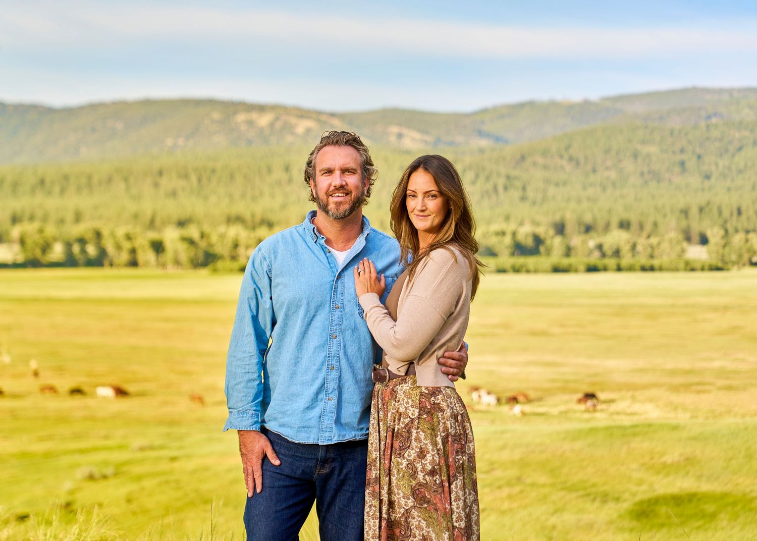 Smiling couple embraces in sunlit field with forested mountains and grazing animals in background.